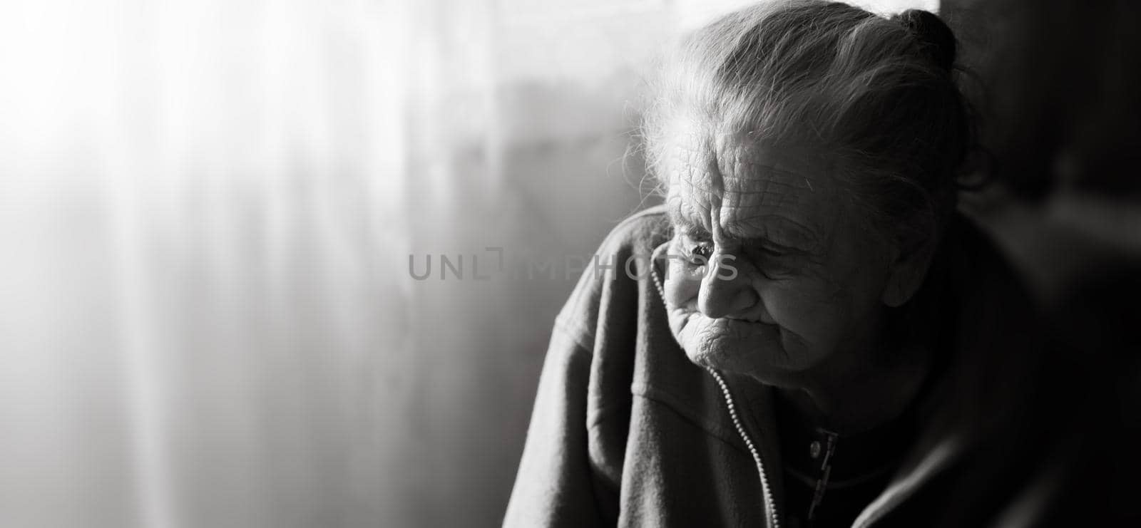 Black and white image of an old depressed woman. An elderly lonely woman sits at a table in the kitchen near the window