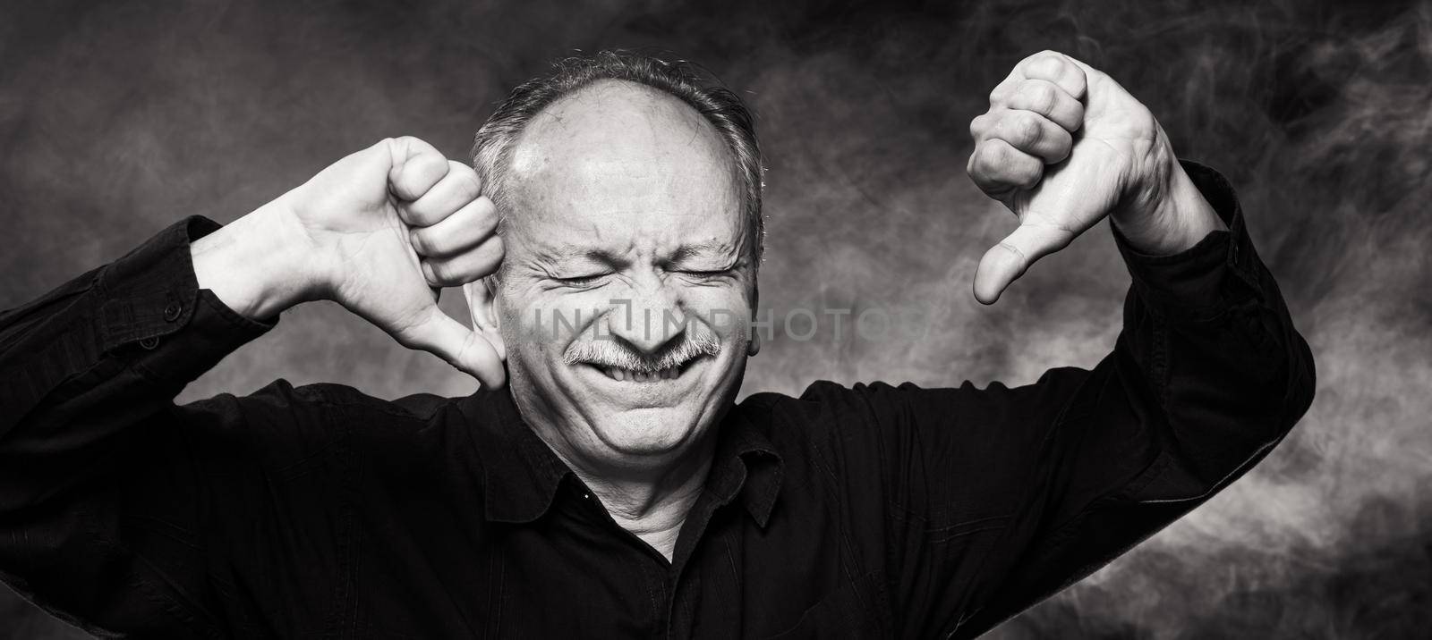 Old macho man gesturing with thumb pointing down. Portrait of an elderly emotional man on a dark background in the studio. Black and white image