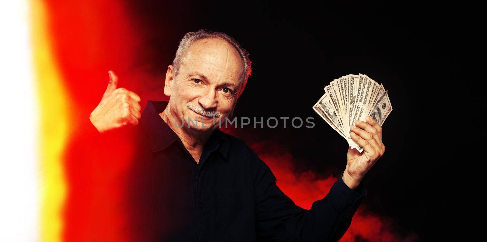 Senior gentleman holding a stack of money. Portrait of an excited old businessman. Happy old man holding dollar banknotes on dark red background with light leak. 