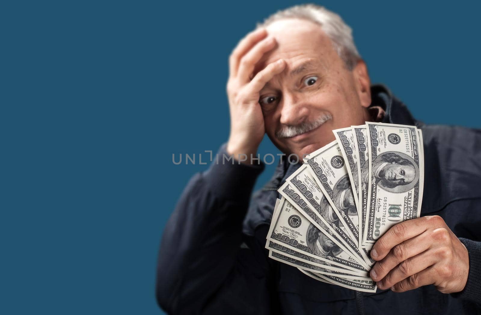 Lucky old man holding with pleasure group of dollar billson blue background with copy-space. Focus on money