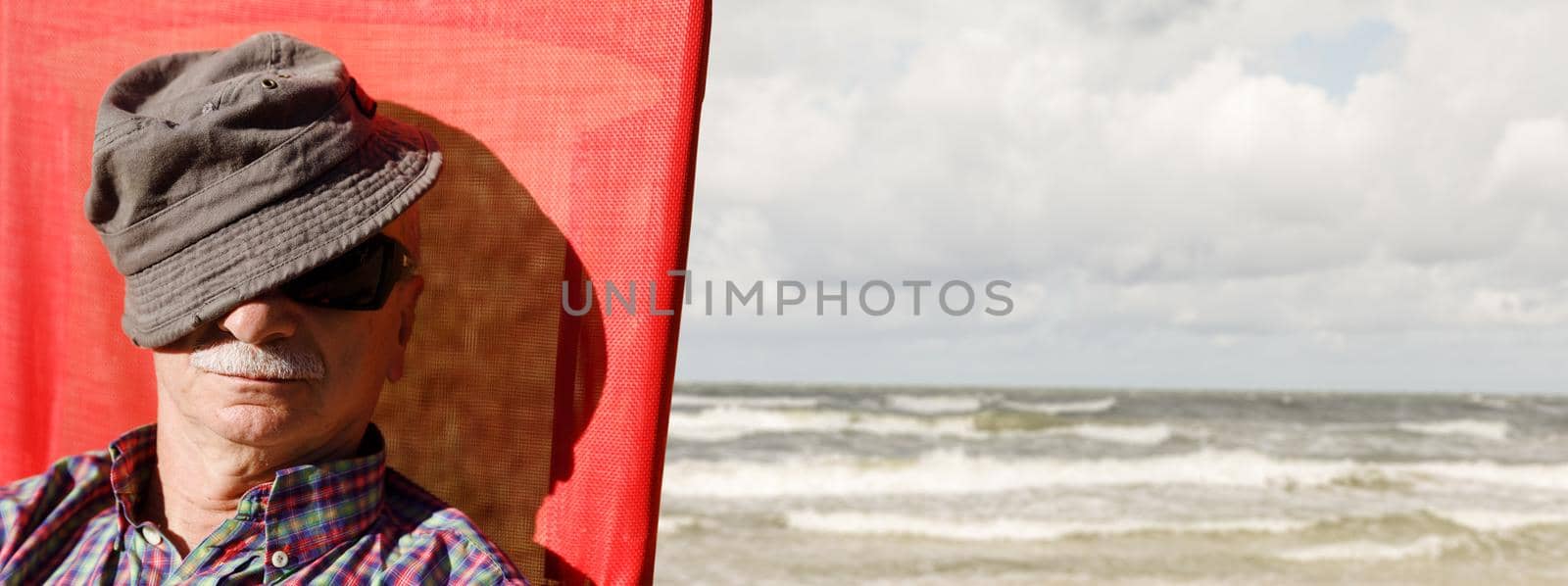 An elderly man sitting on a beach chair on the sea background covering his face by a cap. Lifestyle concept