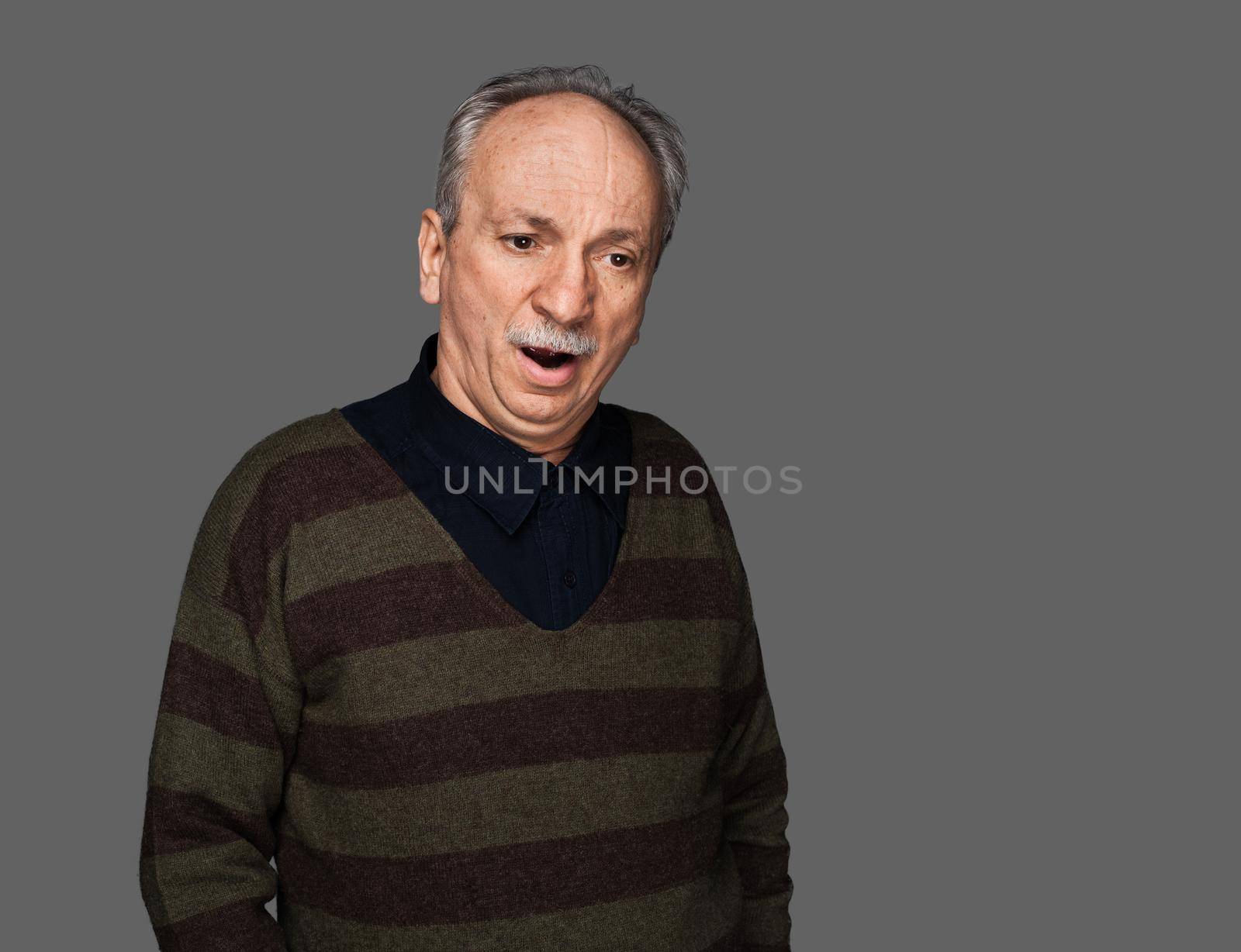 An old man in a sweater yawns. Studio portrait on a gray background