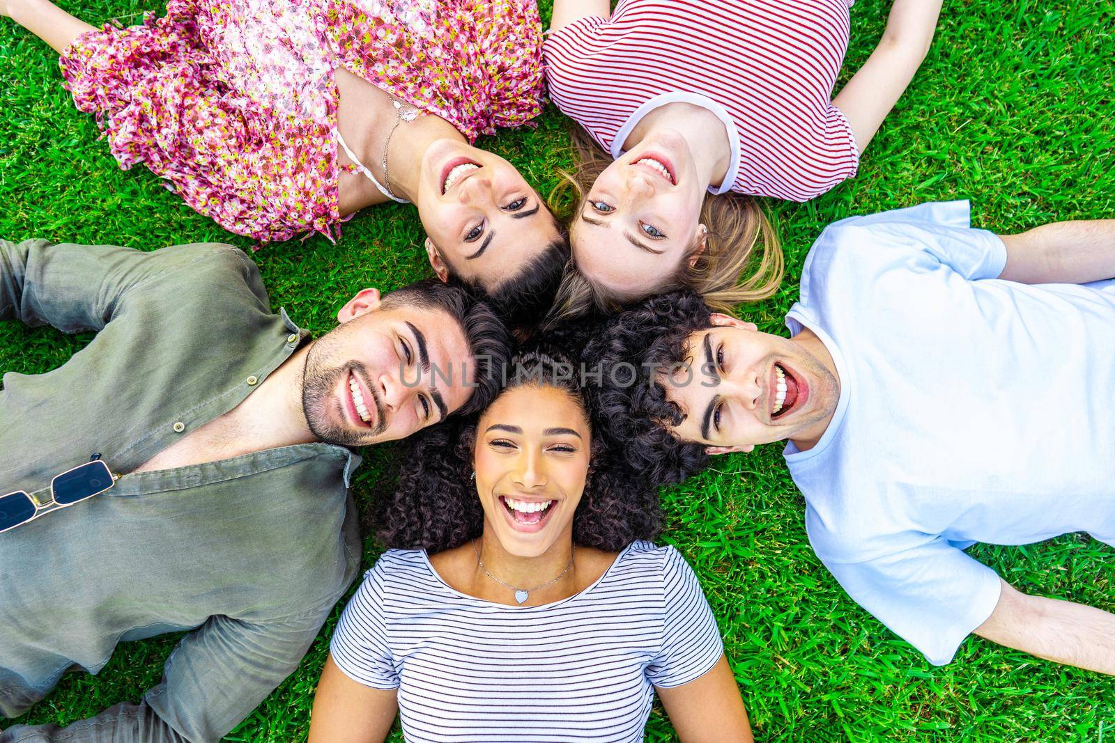 Group of five happy young diverse multiracial gen z friends lying in circle head to head on the grass of a park lawn looking to the camera from the bottom. Concept of friendship without prejudices