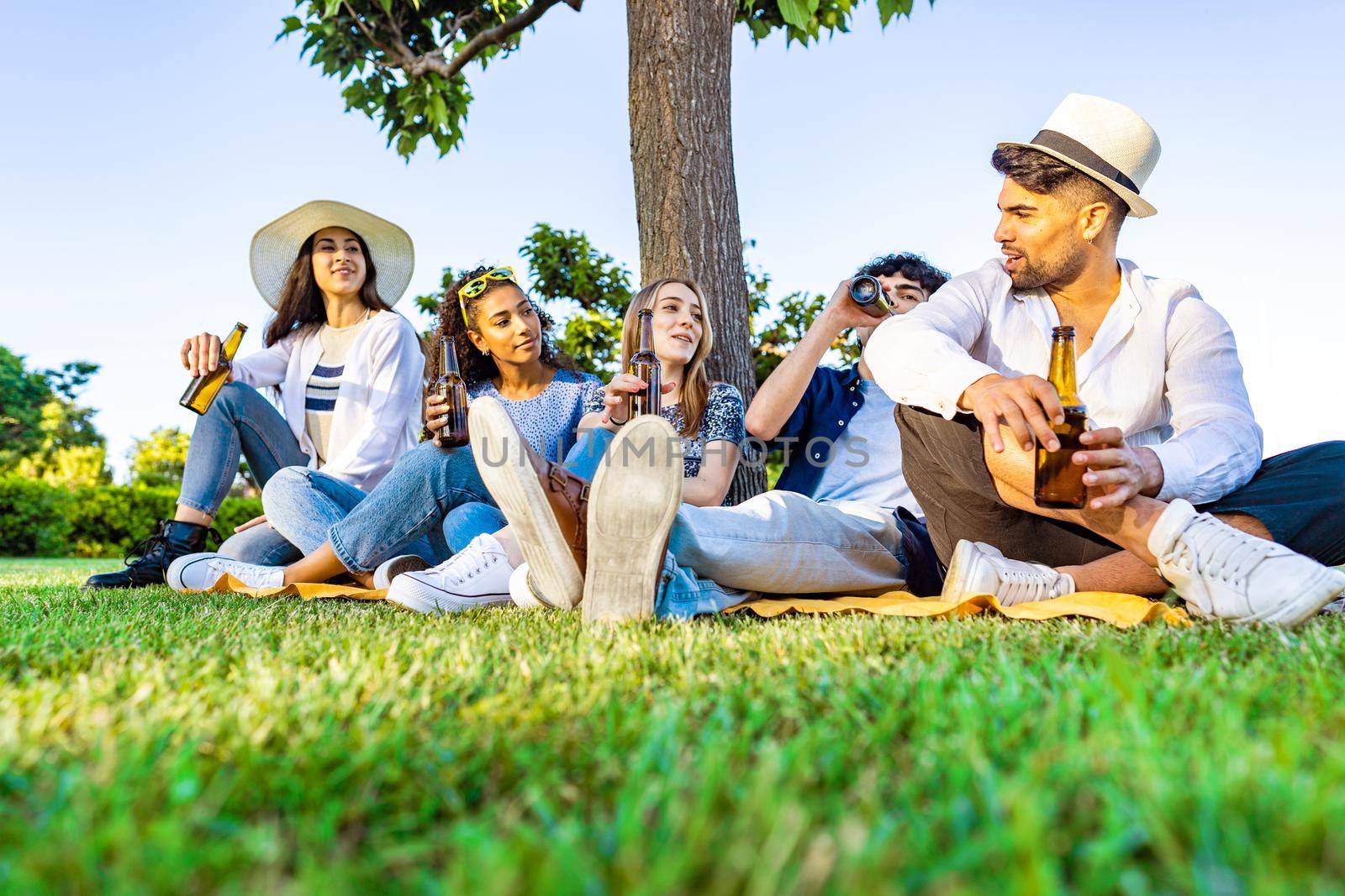 Group of five happy young diverse multiracial gen z people in outdoor party drinking beer from bottle talking each other sitting on grass of city park meadow. Friends having fun with alcohol outdoor