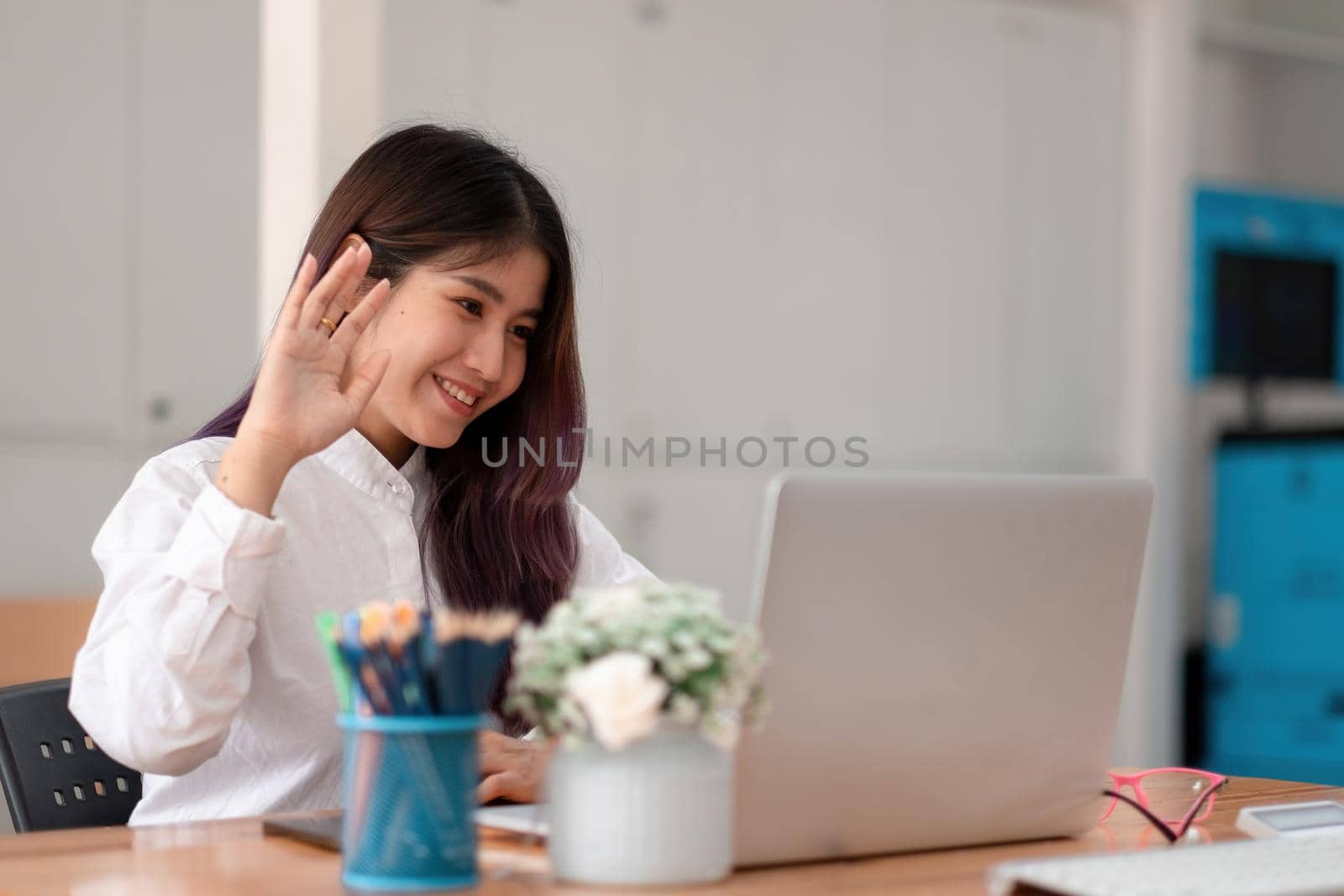 Image of happy asian woman wearing white shirt smiling and waving hand at laptop, while speaking or chatting on video call in office.