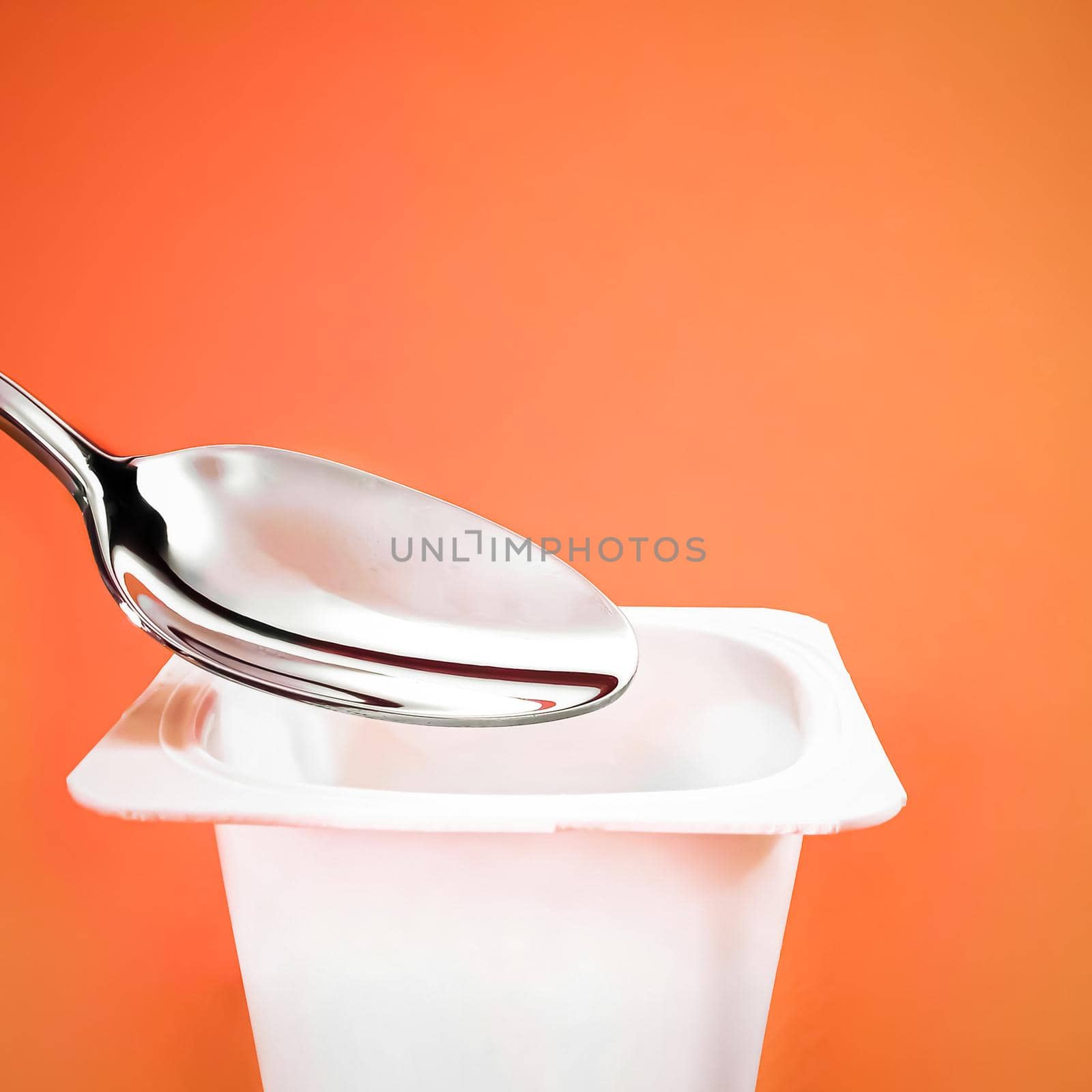 Yogurt cup and silver spoon on orange background, white plastic container with yoghurt cream, fresh dairy product for healthy diet and nutrition balance.