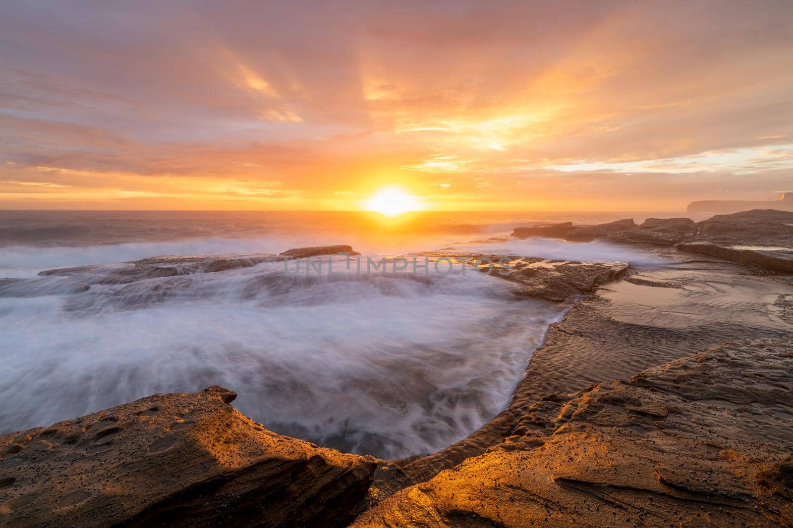 Sunrays burst forth from sunrise sun on beautiful coastline and colouring clouds in teh sky with waves flowing like waterfalls over rocks in foreground.