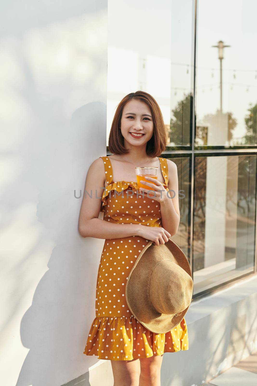 Pretty Asian girl wearing vintage dress and holding hat with orange juice behind wall by makidotvn