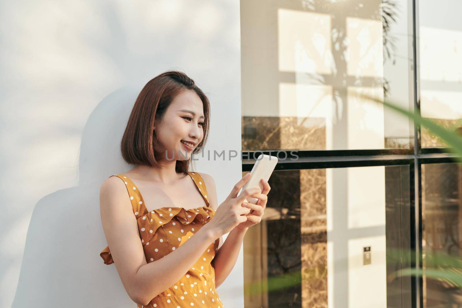 Portrait of young smiling fashion girl standing the wall and using smartphone by makidotvn