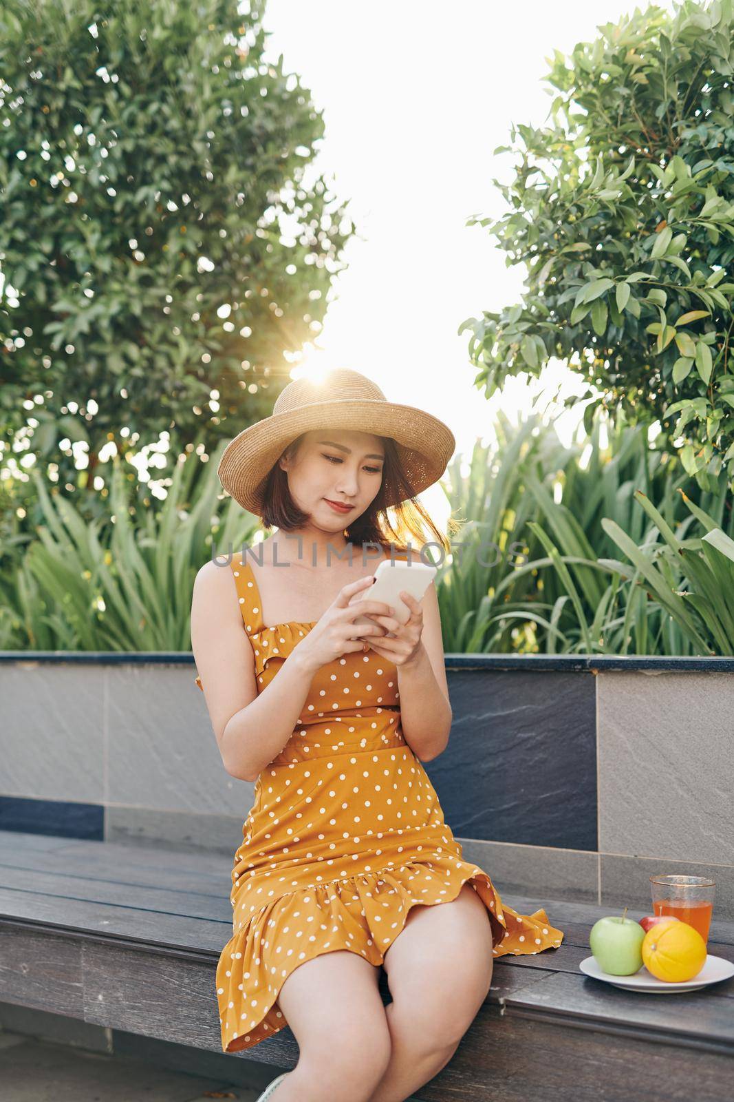 Young Asian girl using smartphone at the park and wearing yellow dress with hat. Summer concept.