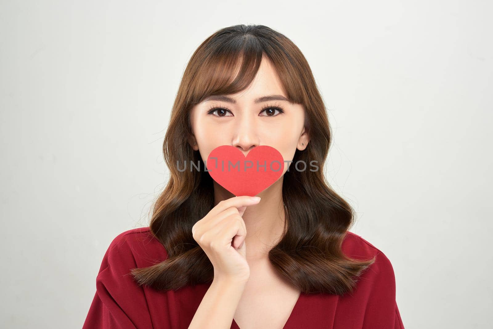 Head and shoulders portrait of attractive Asian woman looking at camera with deep gray eyes while covering her mouth with heart shaped piece of paper by makidotvn