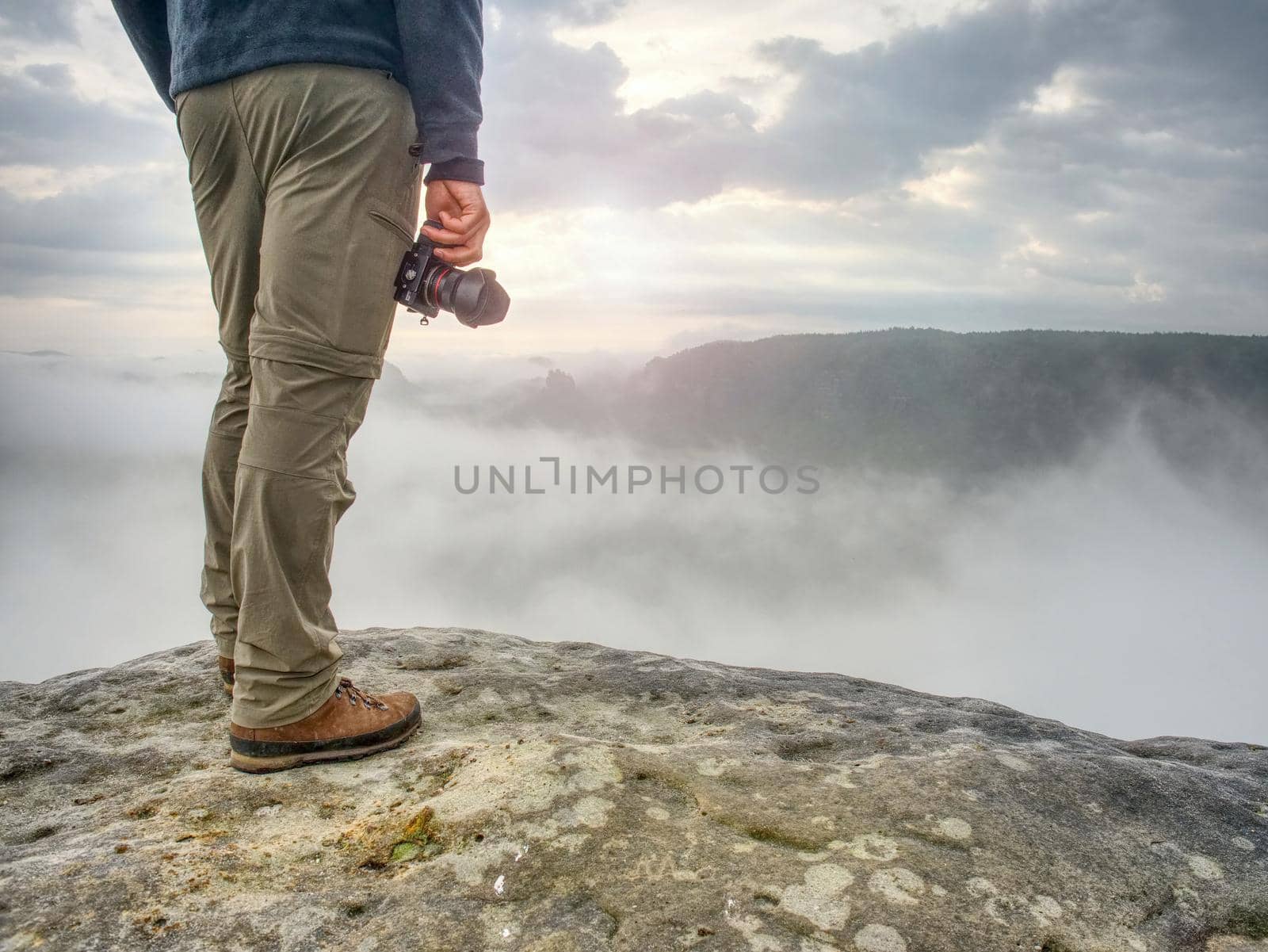 Landscape photograper with camera ready in hand. Man climbed up on exposed rock for fall photos with his digital camera. Cliff above misty autumn landscape.