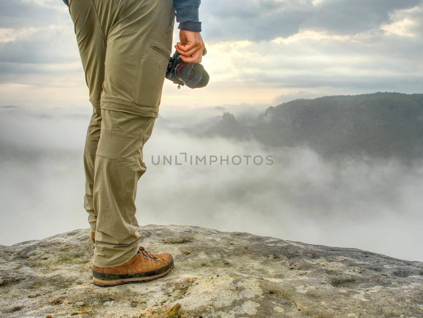 Landscape photograper with camera ready in hand. Man climbed up on exposed rock for fall photos with his digital camera. Cliff above misty autumn landscape.