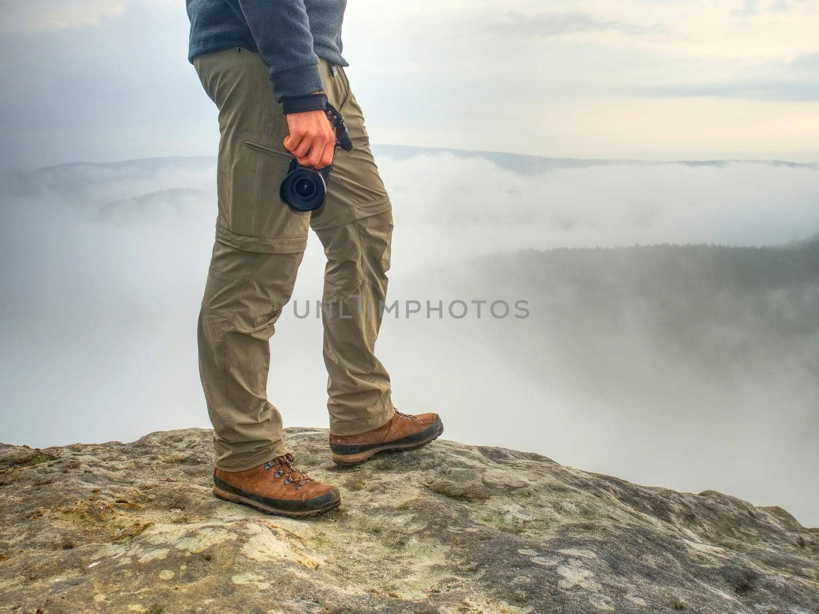 Photographer hand with  camera,  legs, knees and boots. Man takes landscape photos on peak of rock. Dreamy fall  landscape Sun colorized horizon
