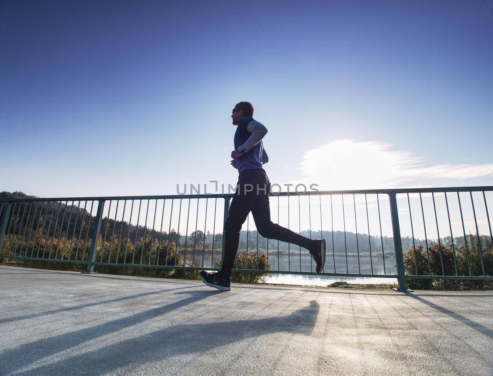 Man is running quickly on the shore bridge. Silhouette of active man exercising and stretching on the lake street. Healthy lifestyle. Alone young fitness man.