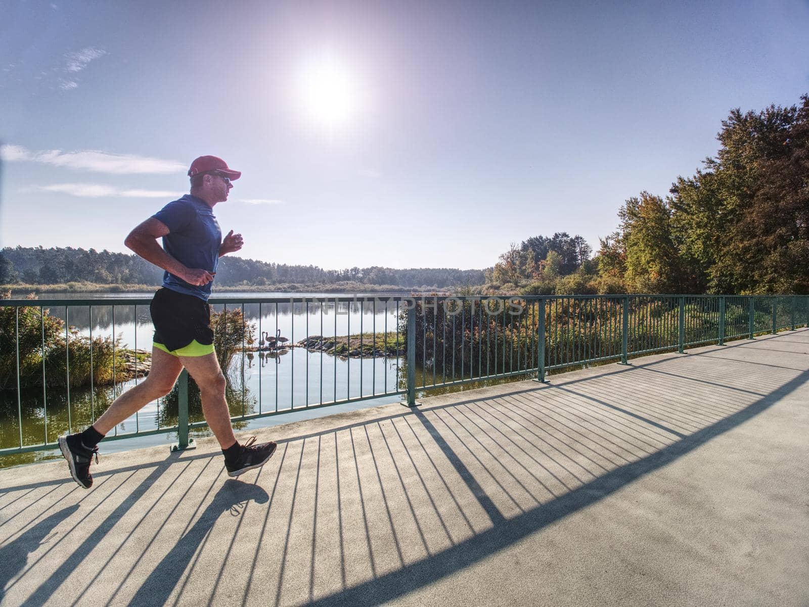 Full section view of fit slim man running on boardwalk. He is in sports clothing. Determined sweaty jogger is exercising by river.