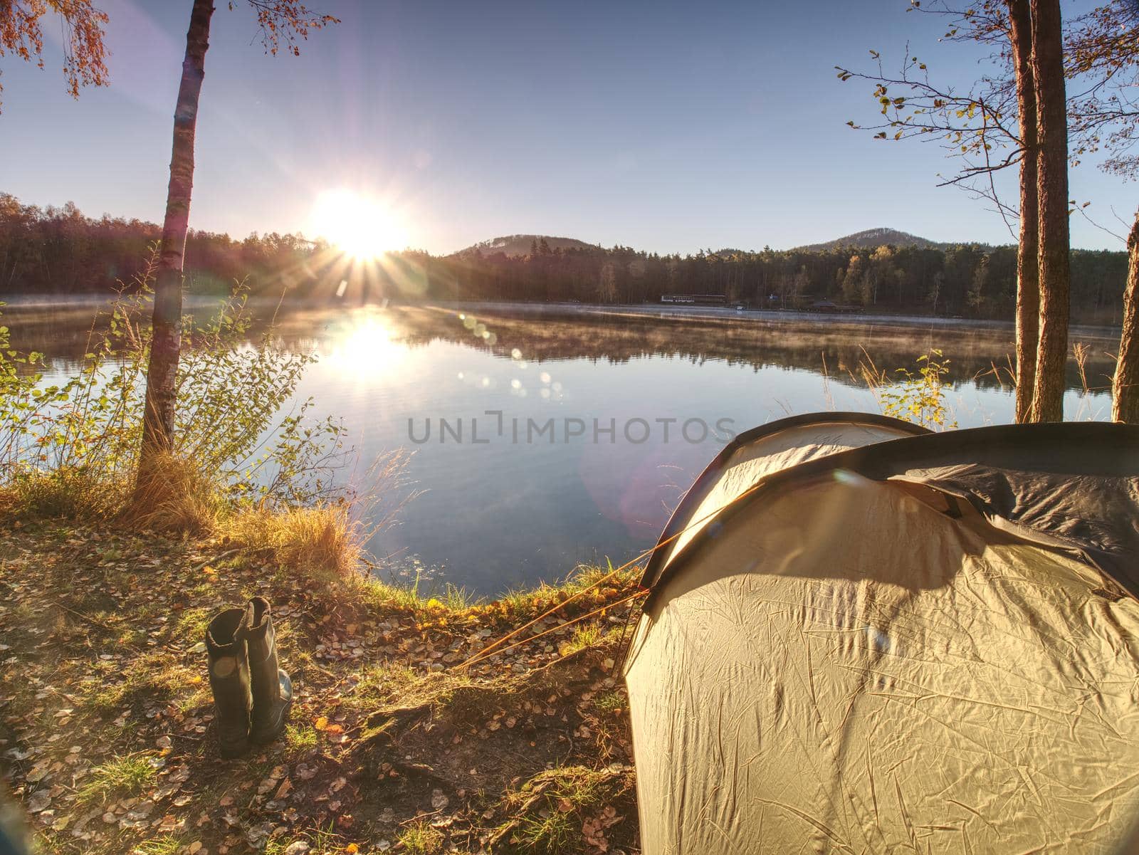 Tent on romantic place at lake shore. Colorful fall forest. Hilly horizon with last sun beams.