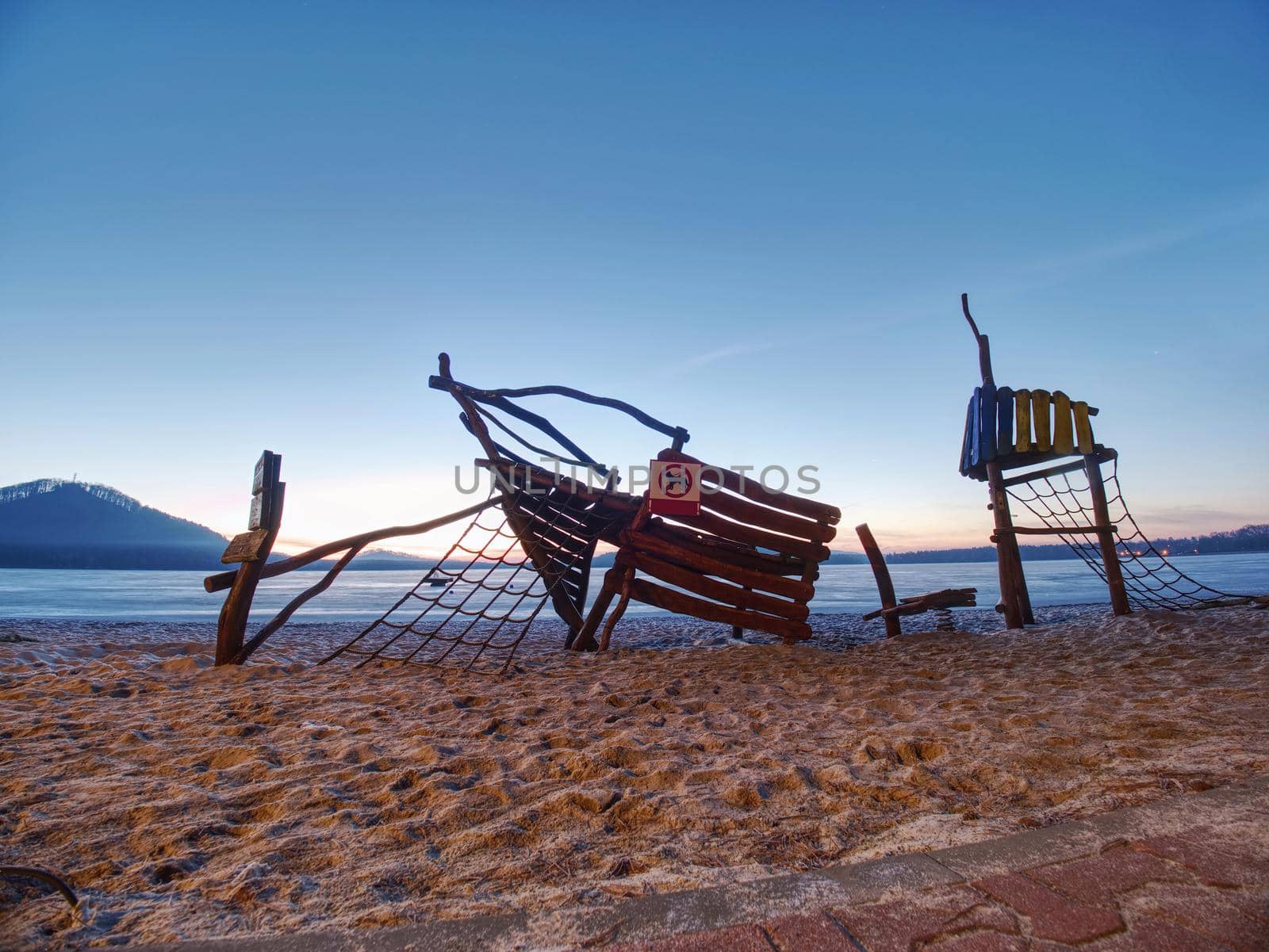 Wooden ship playground attraction for children in swimming bay. Pirate bay with tower built in sand of summer holiday resort. Calm water of lake.