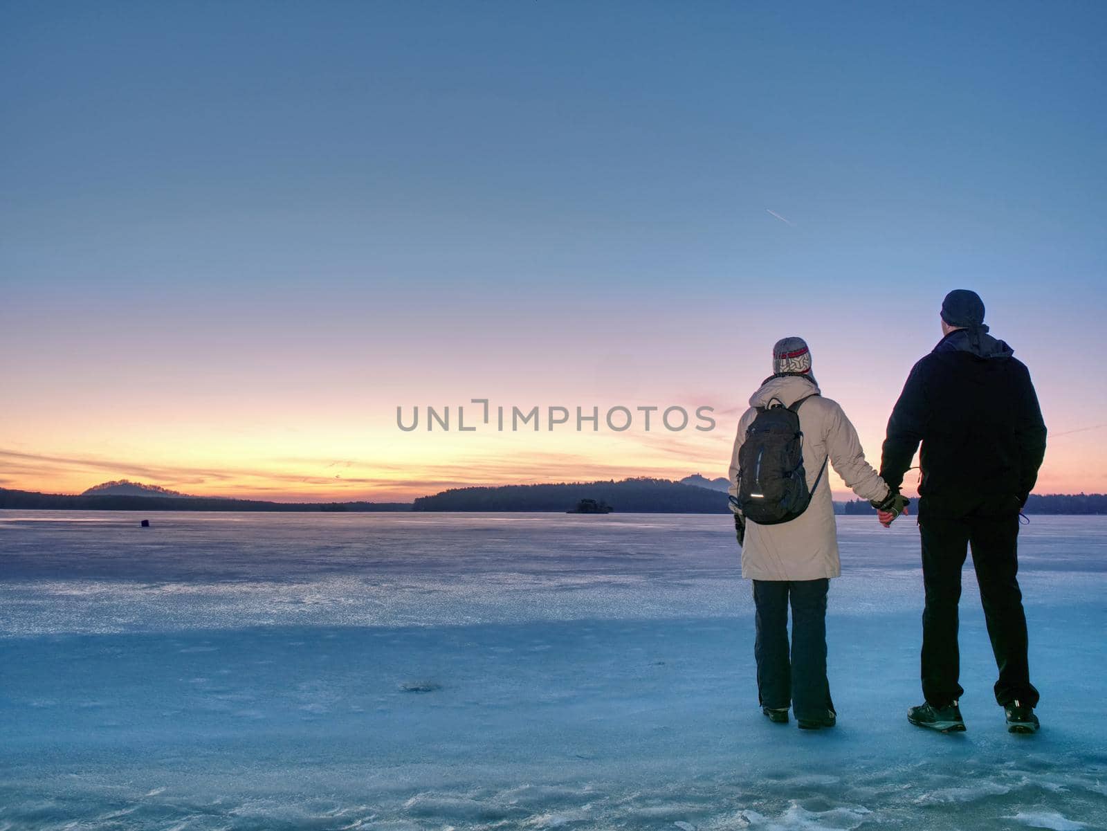 Couple has fun during winter walk on ice of frozen lake. Evening hike at beach of pond. Warm clothes pair with high warm boots.