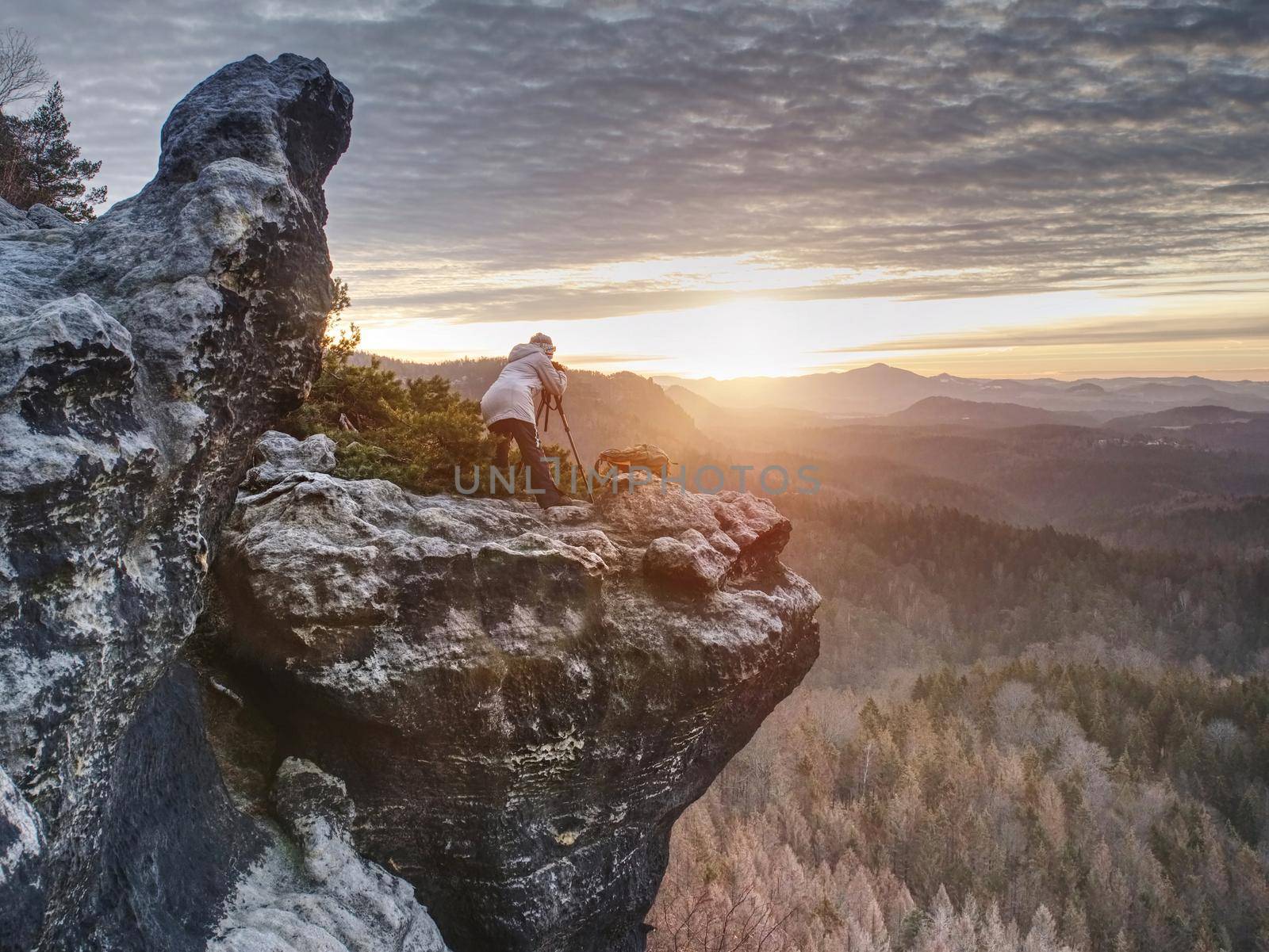 Woman photographer works. Professional artist takes photos with mirror camera and tripod on peak of rock. Dreamy fogy landscape spring orange pink misty sunrise in beautiful valley below.