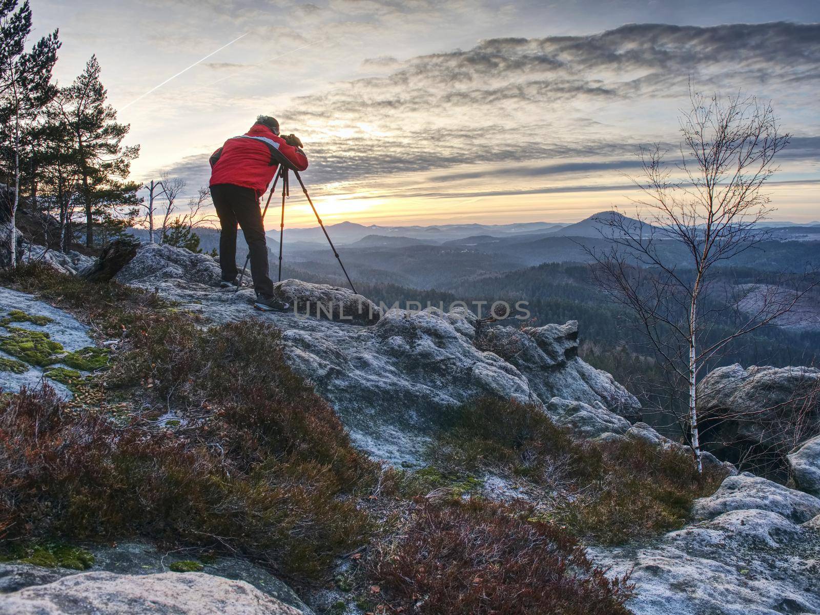 Photographer in red jacket and knitted cap stay with camera on tripod on cliff and thinking. Dreamy misty landscape, misty sunrise