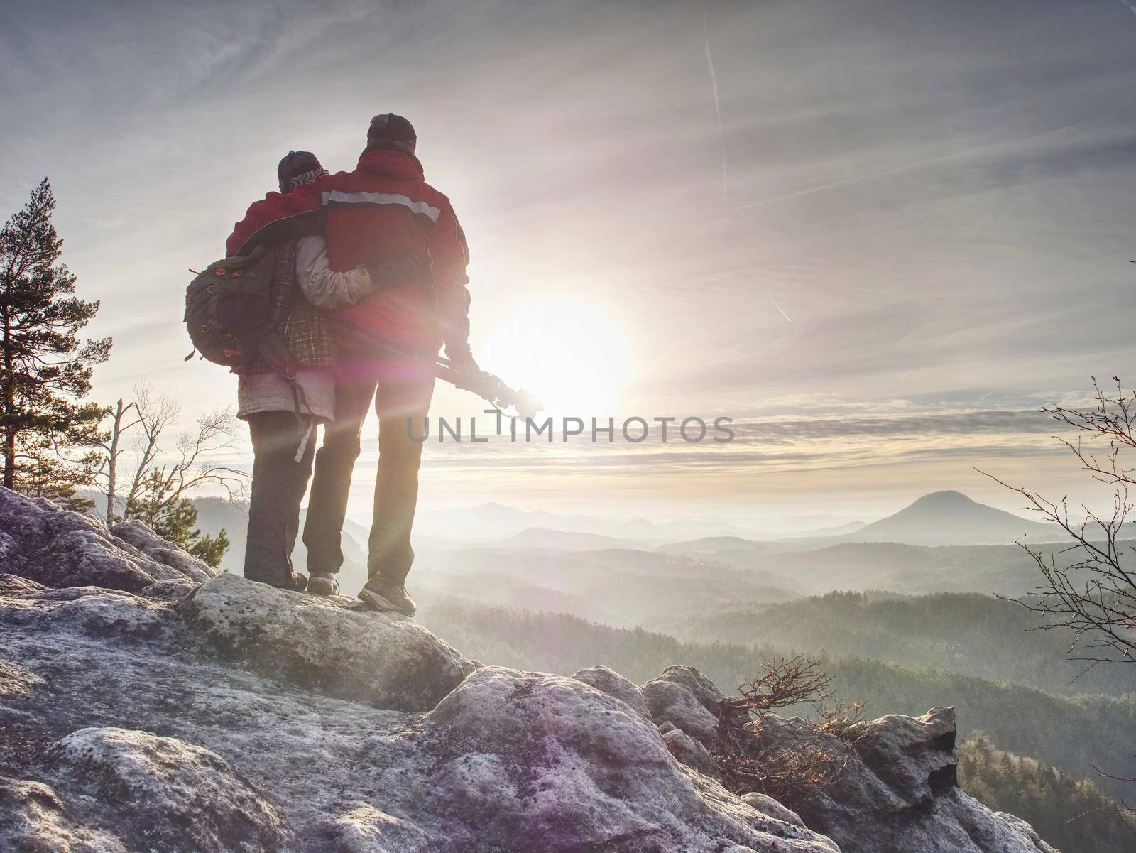 Couple of hikers look down into fogy valley. Photographer stay on cliff and takes photos. Dreamy fogy landscape blue misty sunrise in a beautiful valley below
