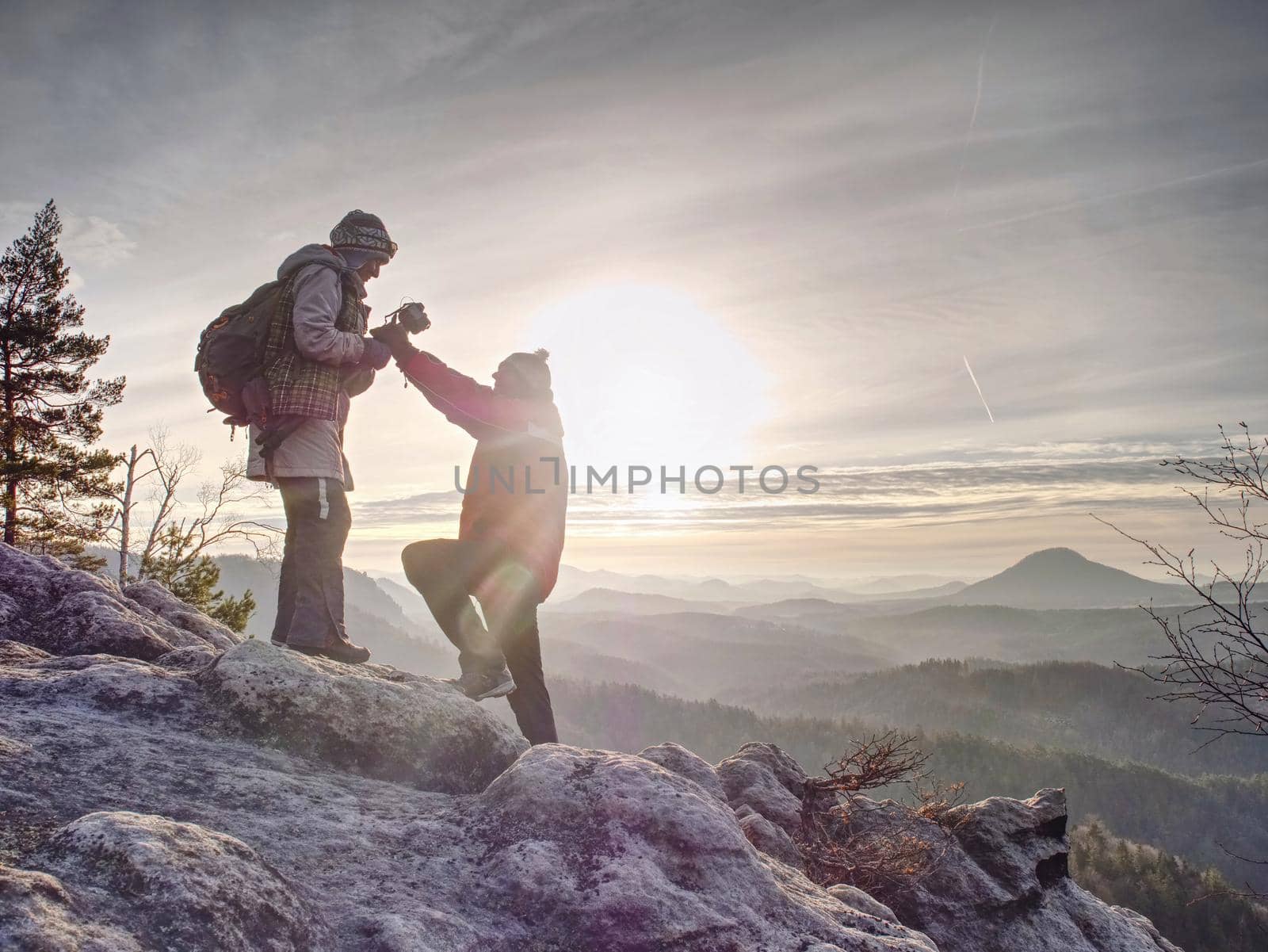 Silhouette man and girlfriend shooting sunset on the top mountain. Photographer with eye at viewfinder of camera on tripod stay on cliff and takes photos talk woman friend