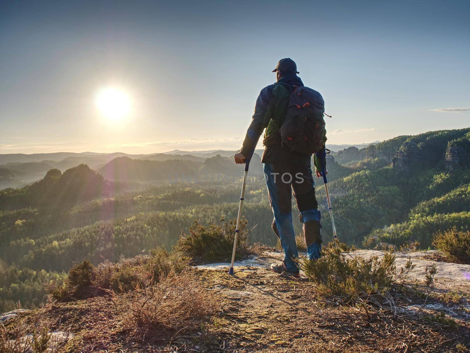 Disabled man with crutches stands on a big rock and looking to mountains at horizon.  Heavy walk in nature trail resort