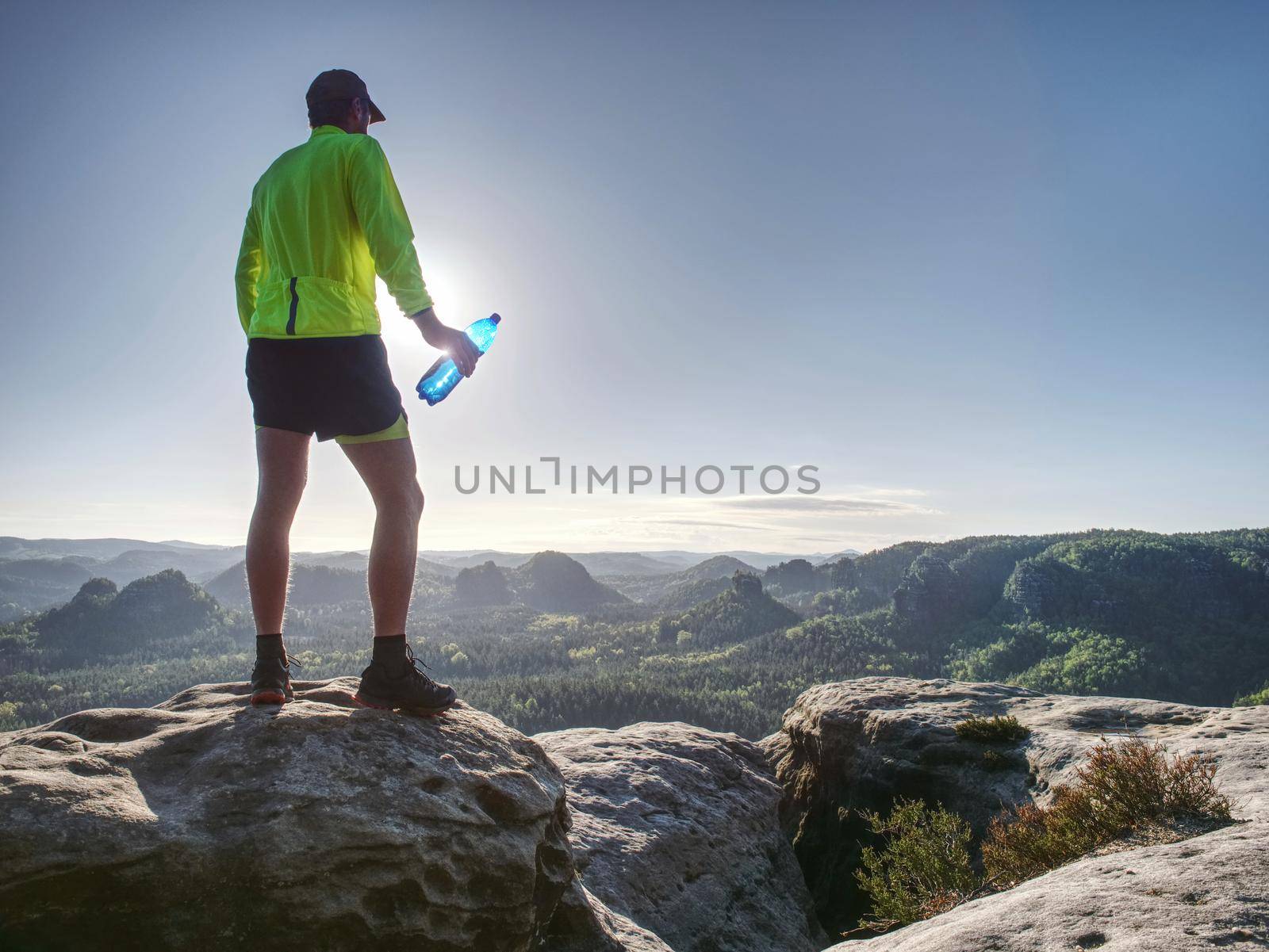 Runner in yellow green shinning jacket and black shorts  jumping on rock. Man athlete while jumping during a trail runnin