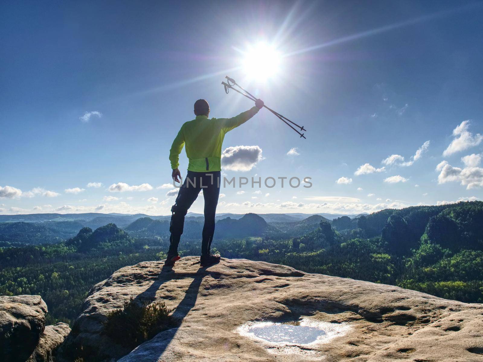 Happy backpacker with trekking poles in the air open mountain valley bellow cliff. Silhouette of tourist with poles in hands