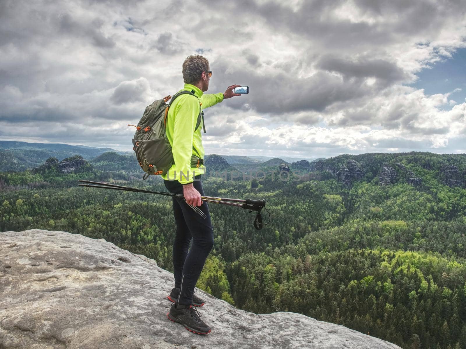 Backpacker with phone in hand. Sunny spring valley in rocky mountains. Hiker with backpack stand on rocky view point