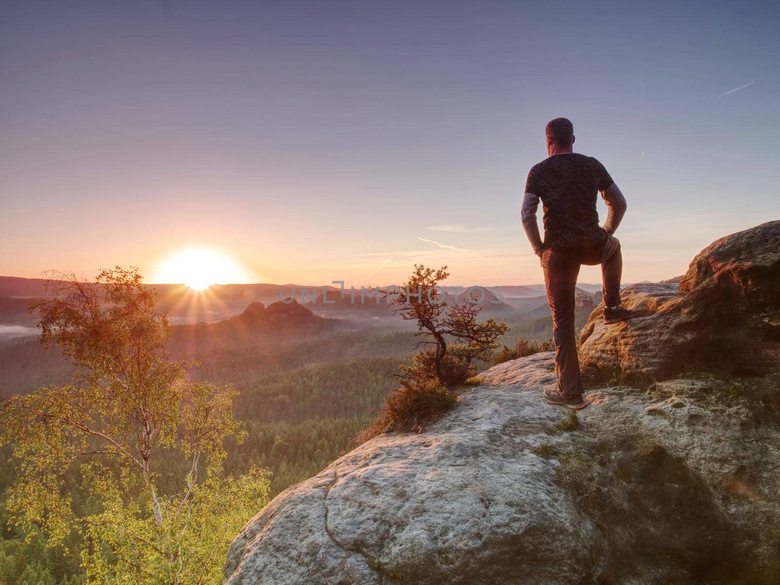 Man on the rock empire with hands in trousers pocket  watch over the misty morning valley to rising sun at horizon.