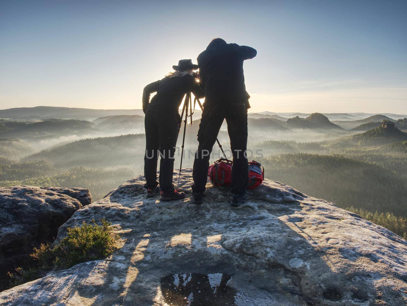 Couple of extreme creative artists work and talk on exposed rock ledge with beautiful view into wild nature. Woman and man takes impressive photos together.