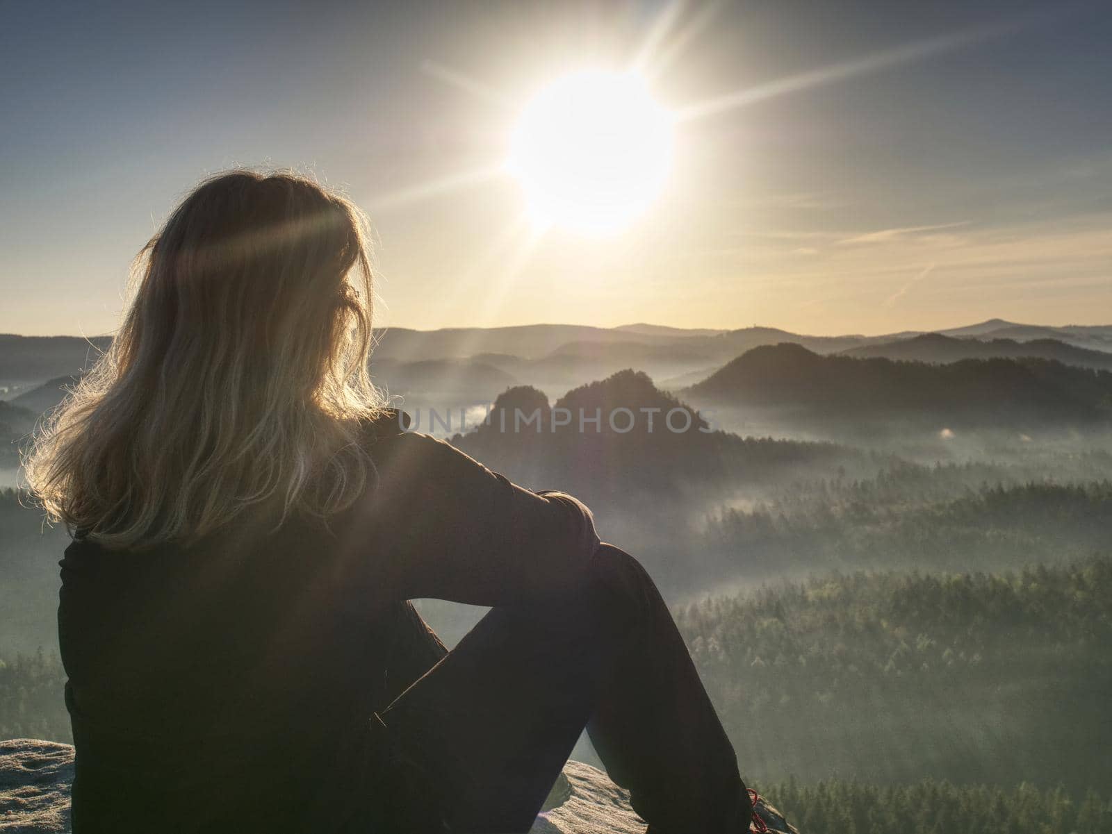 Blond woman sitting on the edge of the mountain cliff against beautiful mountains peak. Travel and lifestyle concept