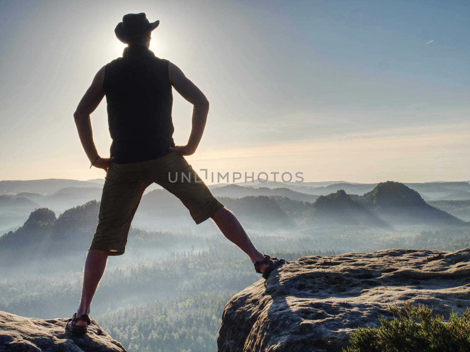 Cowboy style hiker man on cliff of park is watching over misty morning valley to Sun. Man body with akimbo arms