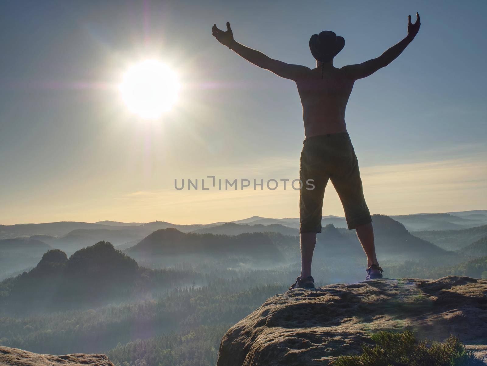 Silhouette of tall naked hiker wearing cowboy hat. Guy looking over large valle from sharp mountain shield.