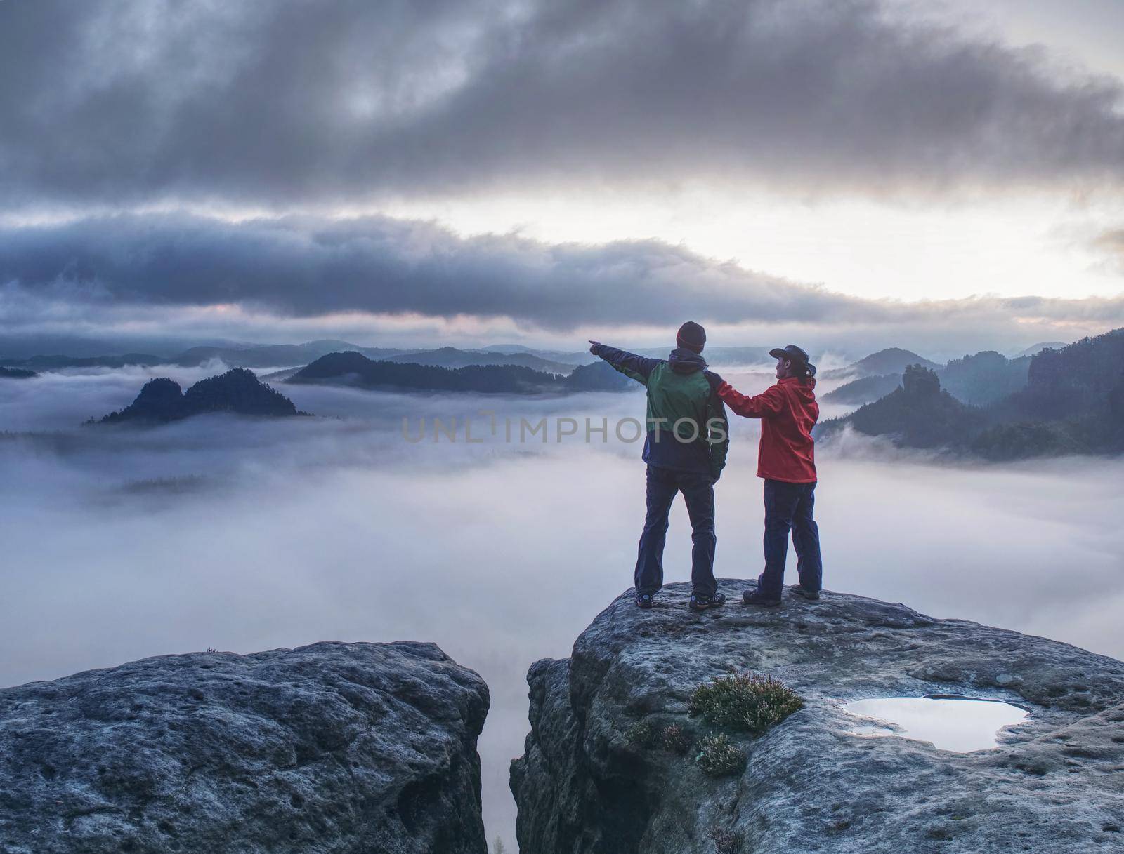 Lovers mirroring in water eye at mountain summit above thick mist.  Climbing couple at top of summit with amazing aerial view