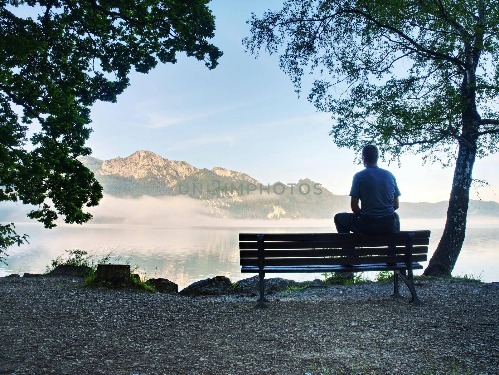 Man sit lone on bench in park next lake. Mountain range by rdonar2