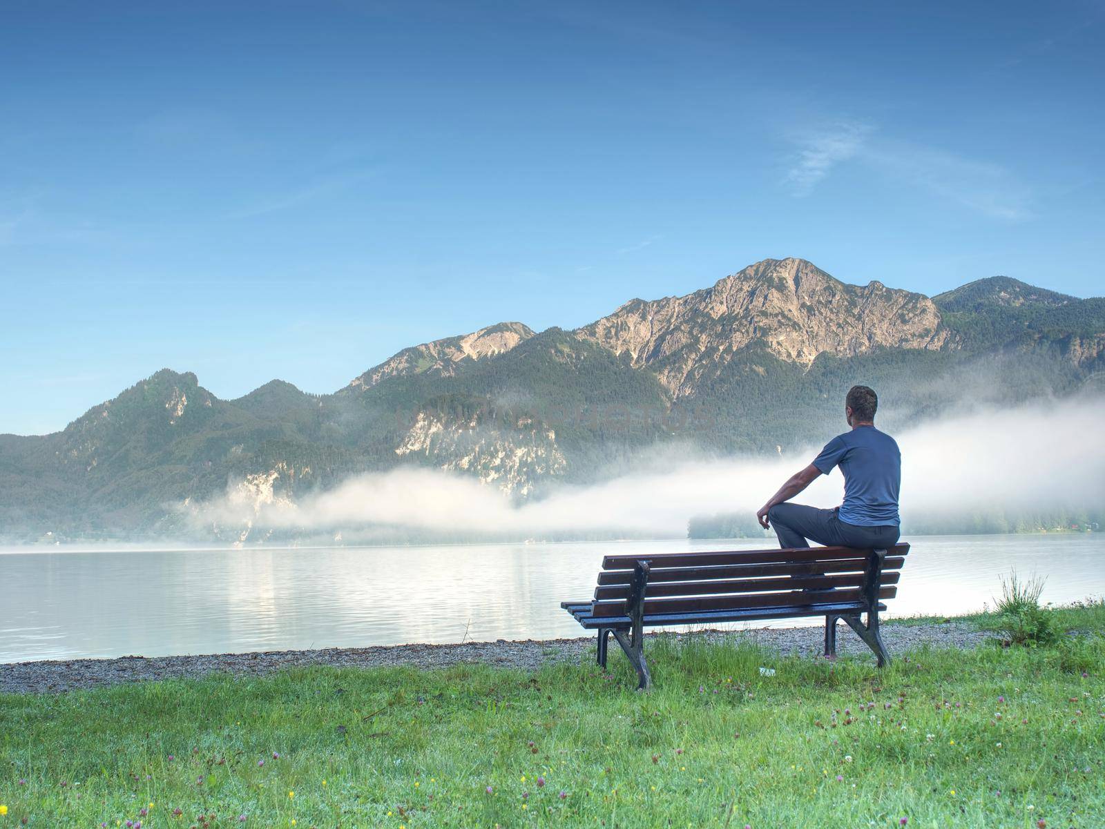 Man sit lone on a bench in park next lake. Mountain range with amazing view over smooth lake surface.