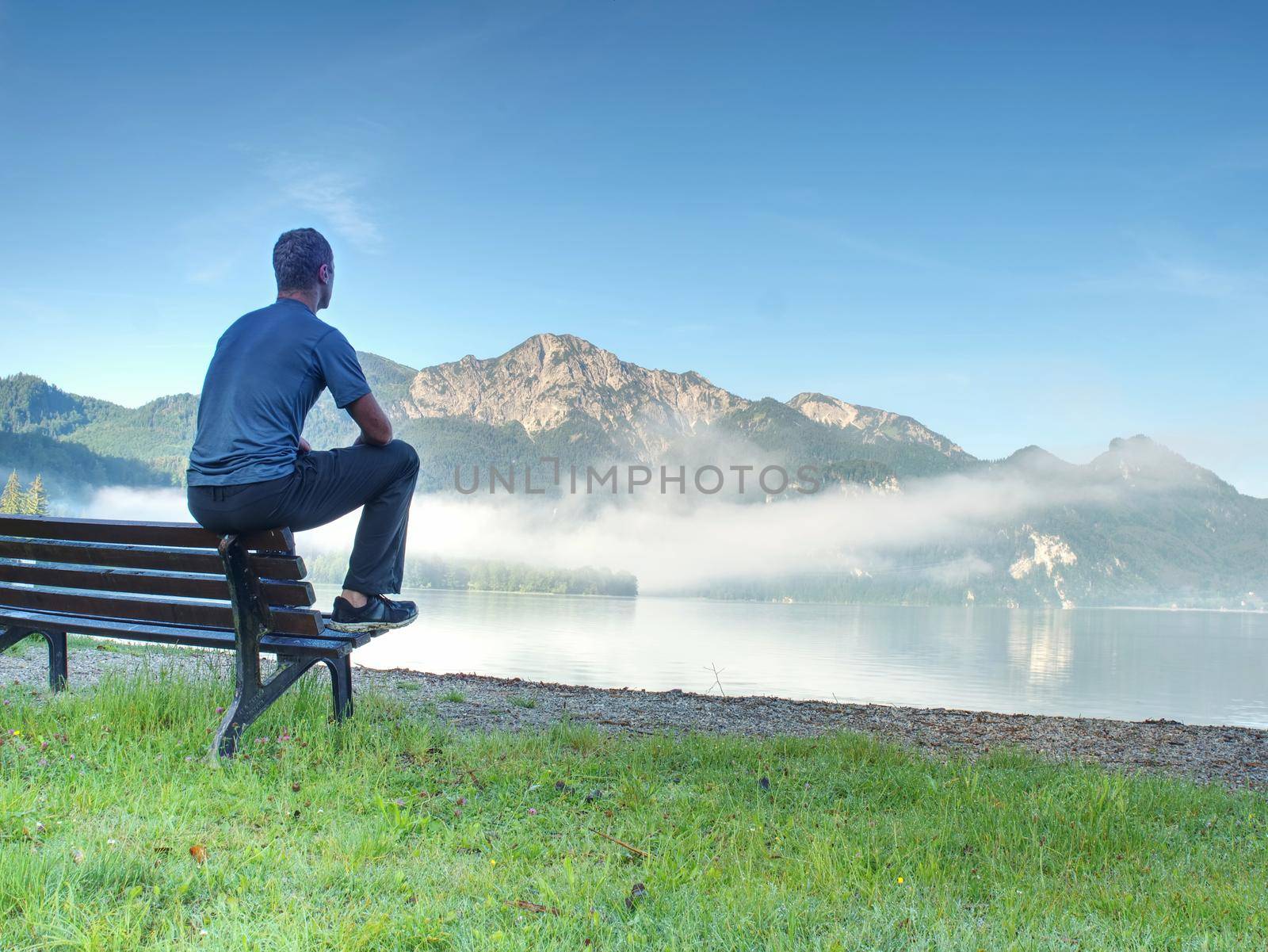 Man sit on wooden bench at coast of picturesque lake bellow blue mountain summits. Calmness, silence and harmony concept.