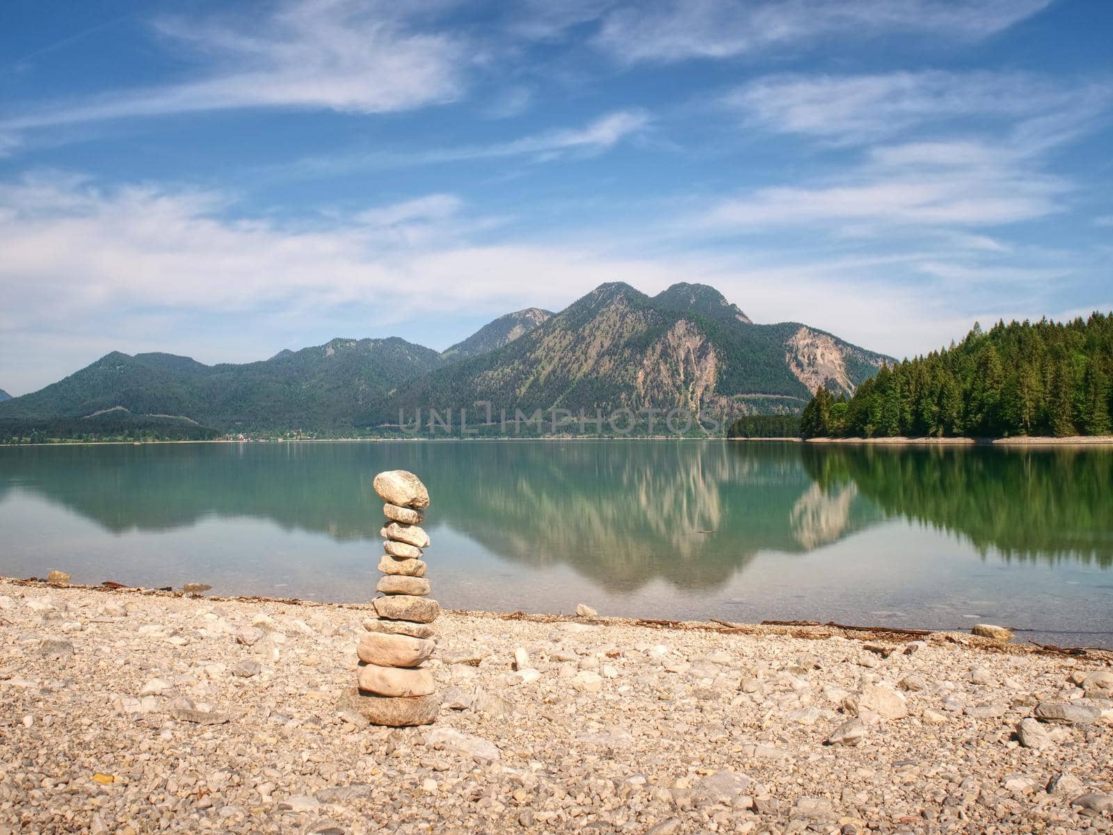 Pyramid of flat stones on a pebbly lake beach, the mountains mirroring in smooth water level.