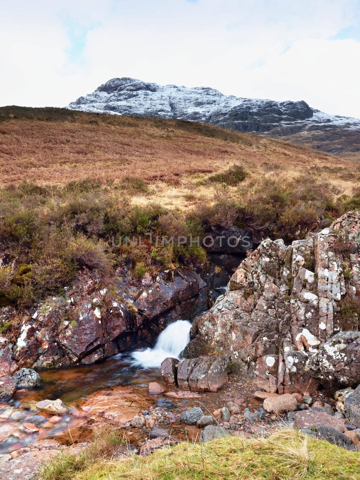Rapids in small waterfall on stream, Higland in Scotland an early spring day. Snowy cone of mountain in clouds. Dry grass and heather bushes on banks