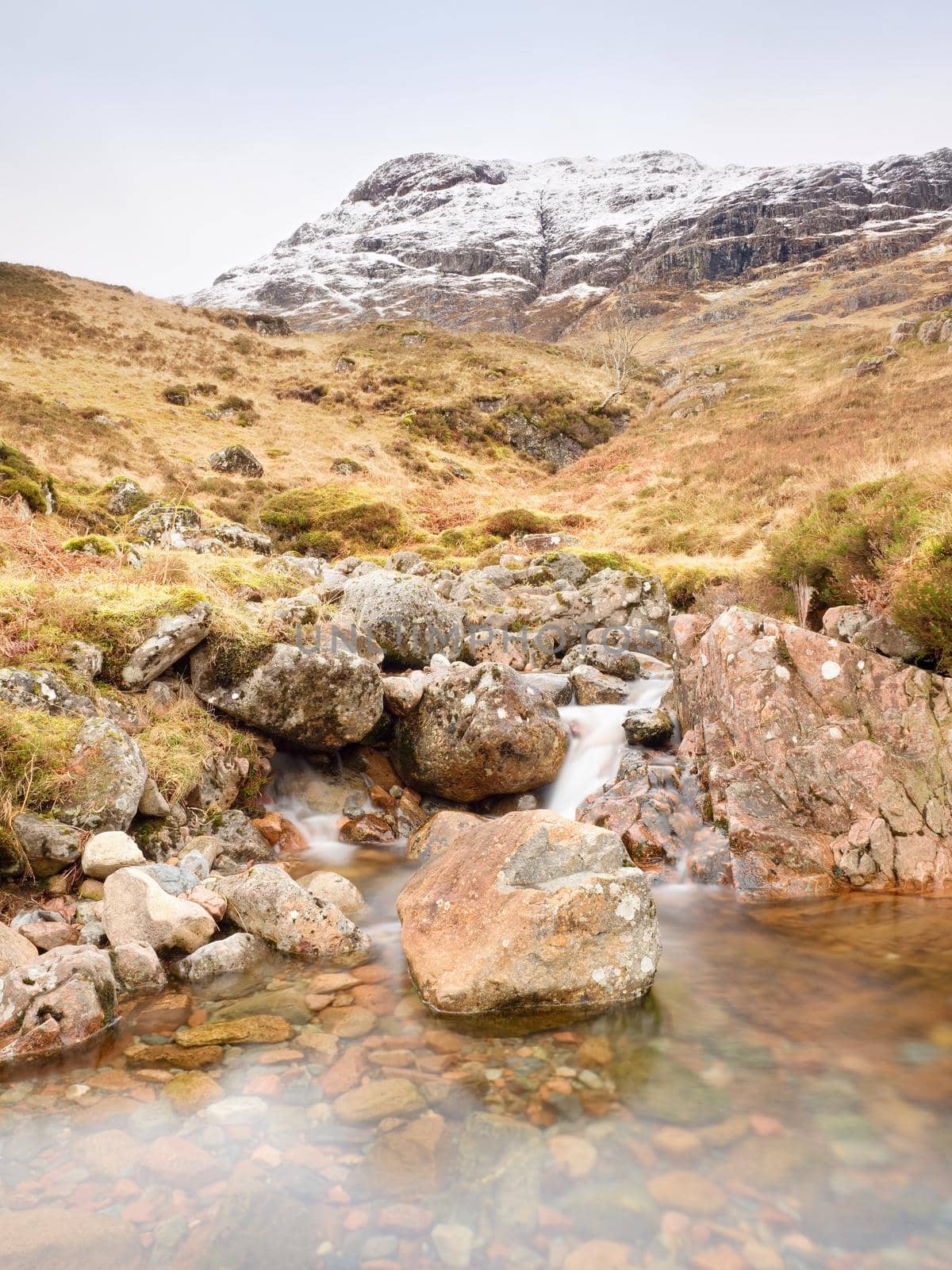 Rapids in small waterfall on stream, Higland in Scotland an early spring day. Snowy cone of mountain in clouds. Dry grass and heather bushes on banks
