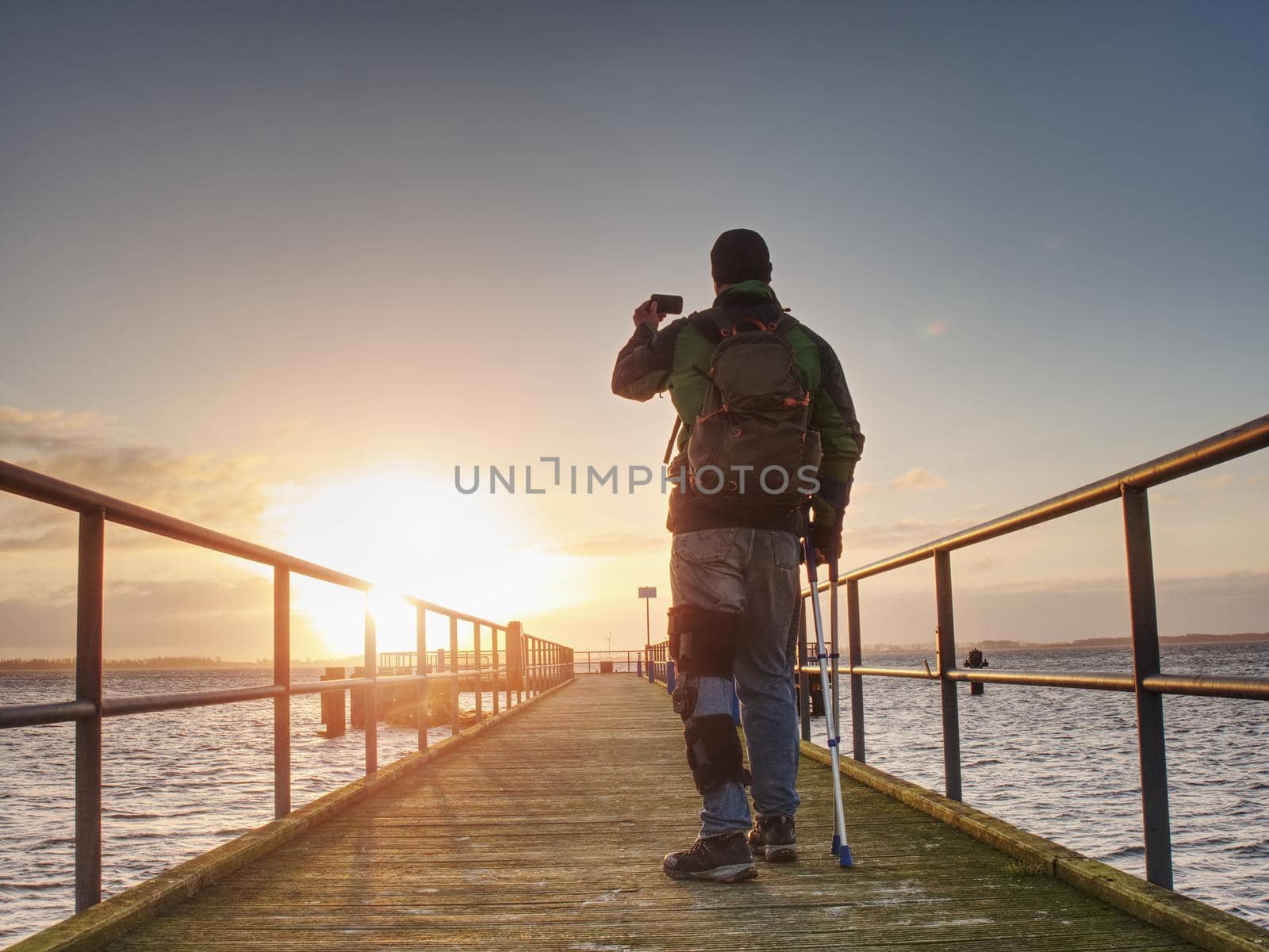 Man with phone in hand takes photo of sunrise on ferry port mole.  Hurt man with crutches.