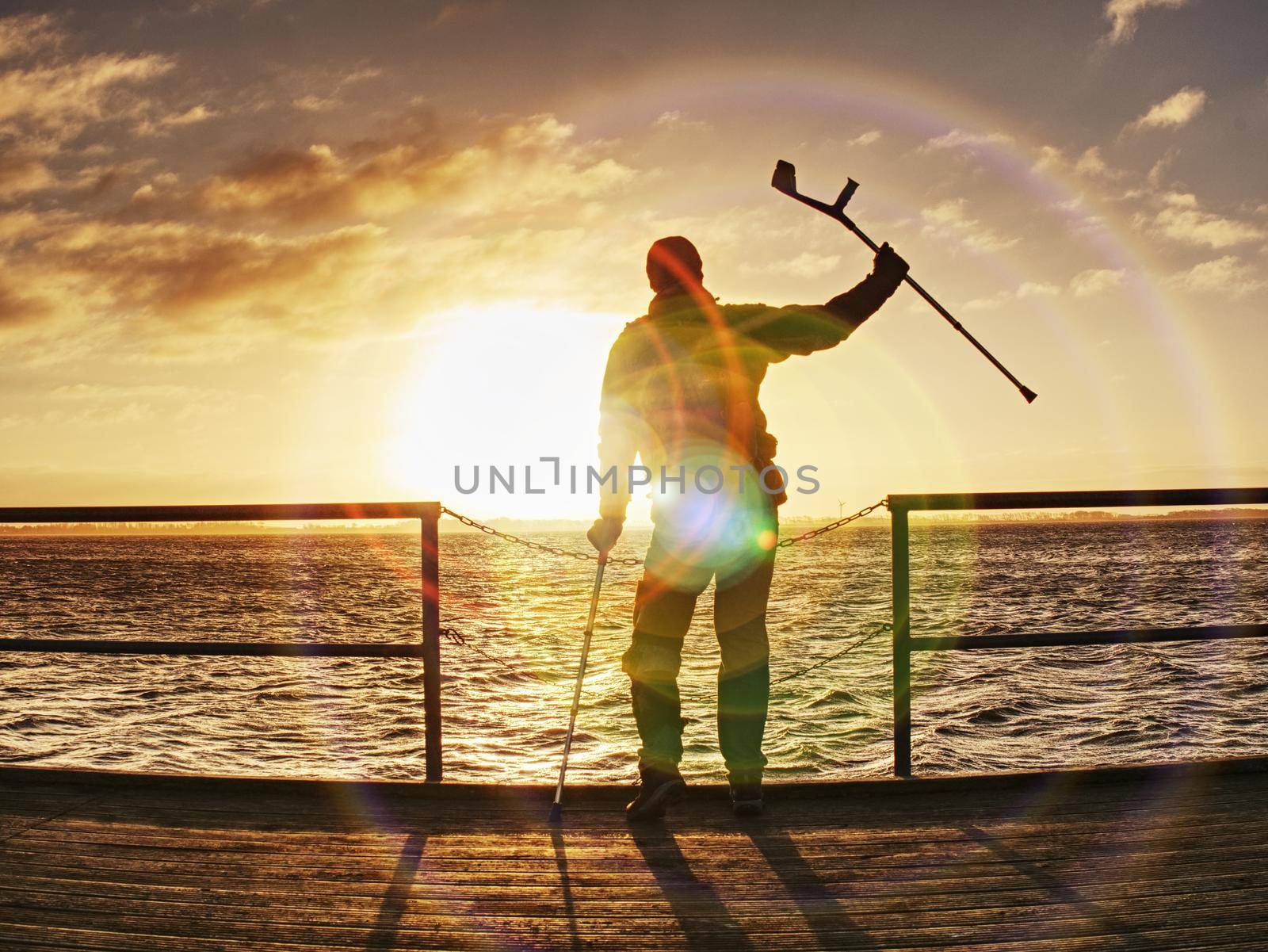 Happy disabled man on bridge holding his forearm crutches above head. Freedom gesture. Fantastic morning with clear sky and  smooth water level