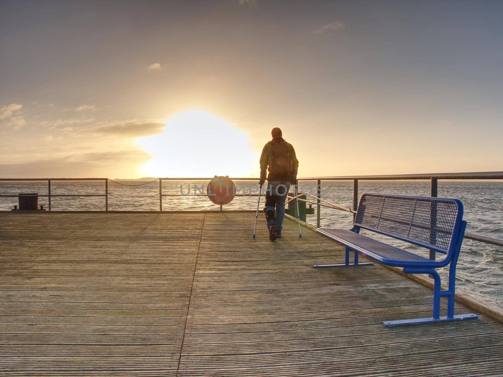 Traveler with backpack walk dificult with medicine poles.  Young tourist on bridge  near sea at the sunset