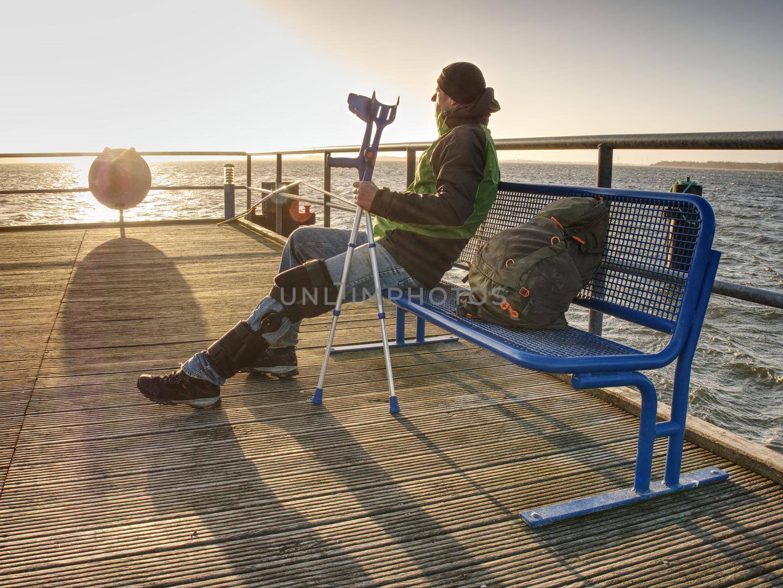 Hurt man having rest, looking into distance and meditating. Person watching sunset sitting against blue sea 