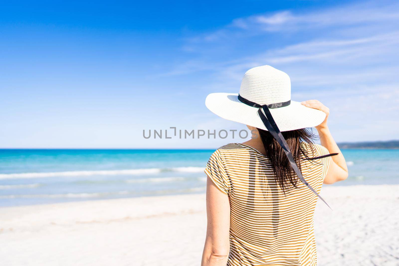 Tropical sea travel ADV banner style: unrecognizable woman holding white large hat with a hand looking to the blue ocean water standing on white silked sand under blue sky. Escape from everyday life by robbyfontanesi