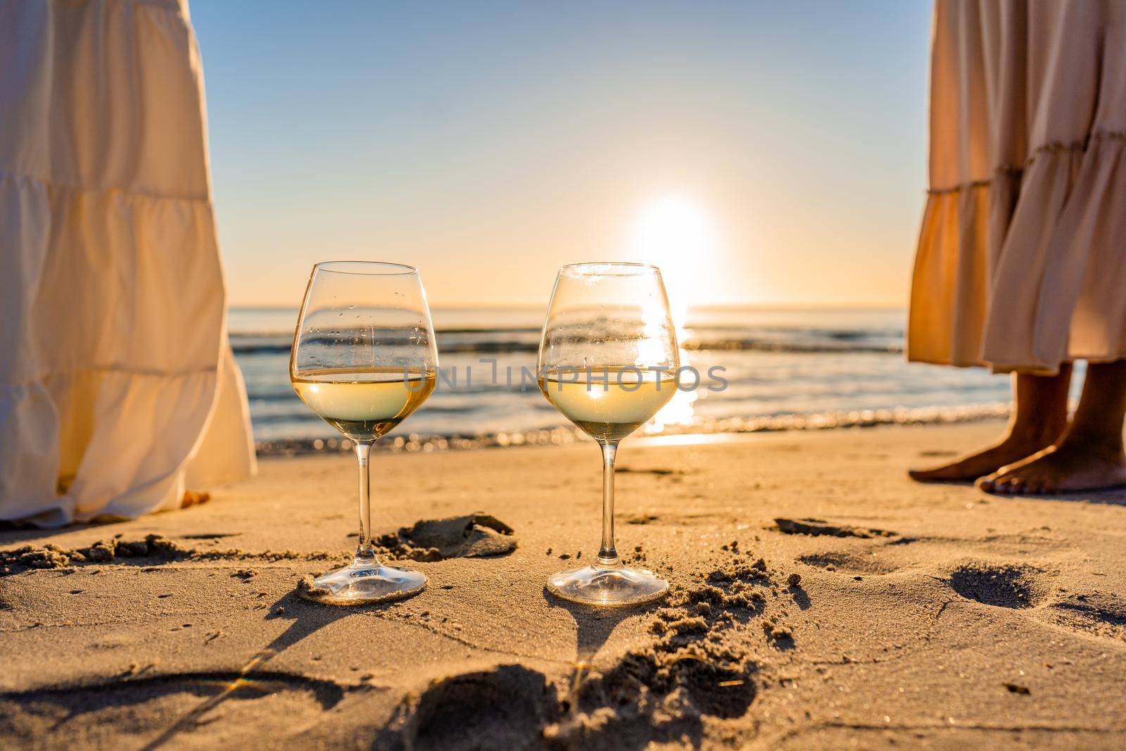 Sunset or sunrise beach party scene with two unrecognizable multiracial boho girls wearing long white dress barefoot on the ocean beach sand. Wine glasses on the ground with sun reflections effect by robbyfontanesi