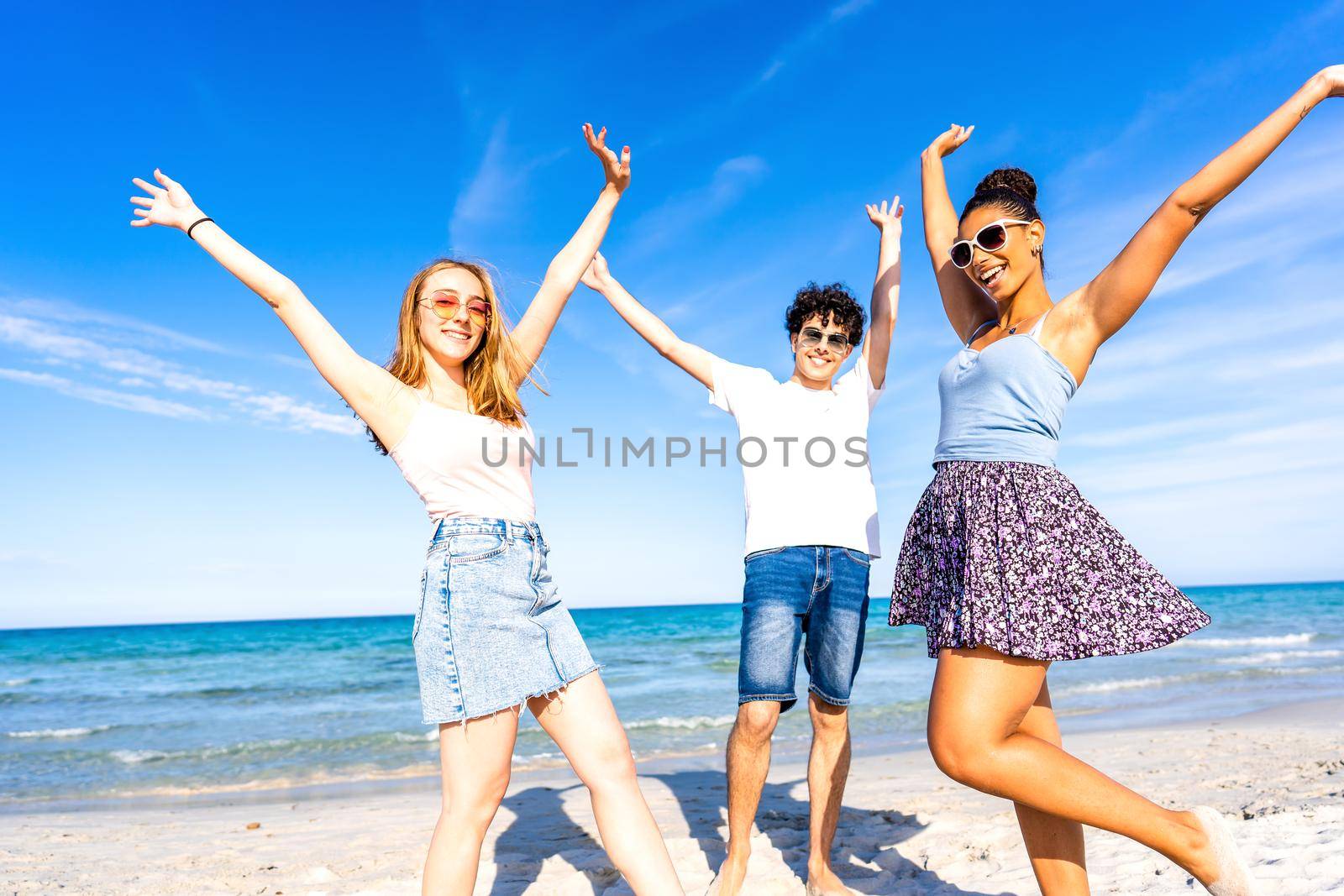 Three very happy friends having fun at the beach posing laughing looking at camera for a portrait with raised arms. Gen z multiracial carefree people in tropical beach with white sand and blue sky